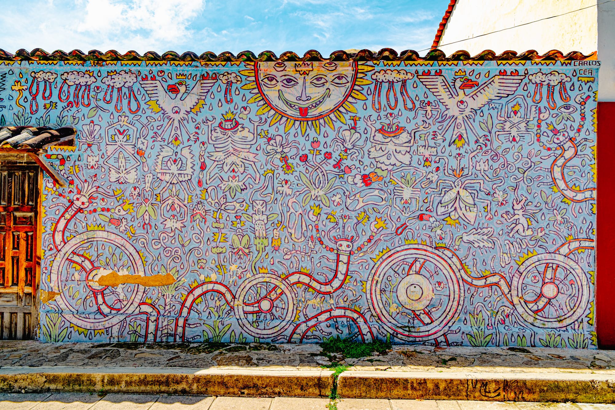 A vibrant mural by Carlos Cea in El Cerrillo, San Cristóbal de las Casas, featuring a whimsical sun with multiple eyes above a dense field of abstract red and yellow linework filled with symbols, creatures, and wheels, reflecting cosmic energy and playful mysticism