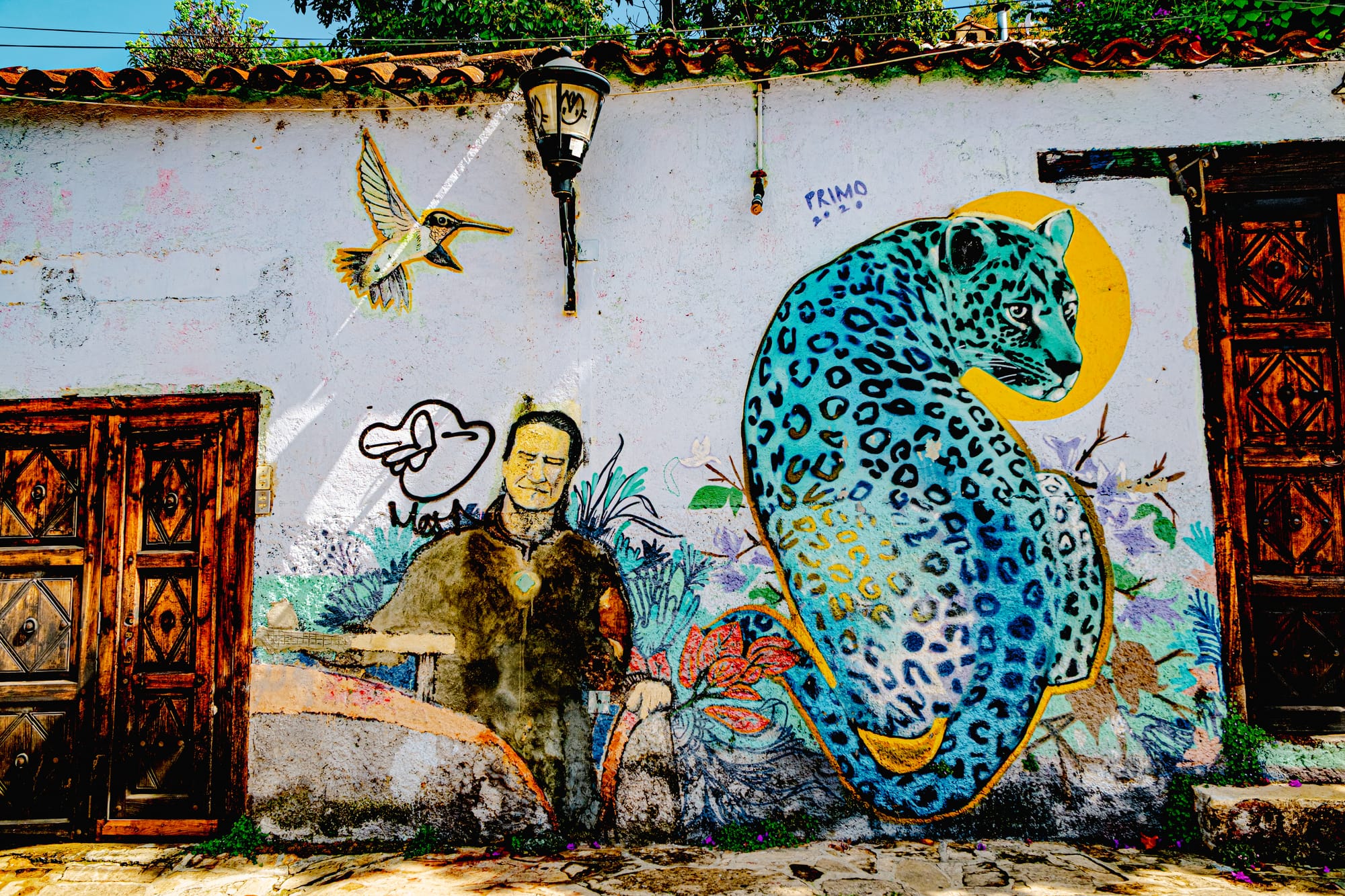 A mural in El Cerrillo, San Cristóbal de las Casas, showing a turquoise jaguar with golden accents beside a seated man and a hummingbird, painted on a white adobe wall with wooden doors, symbolizing harmony between humans, animals, and nature in Chiapas street art.