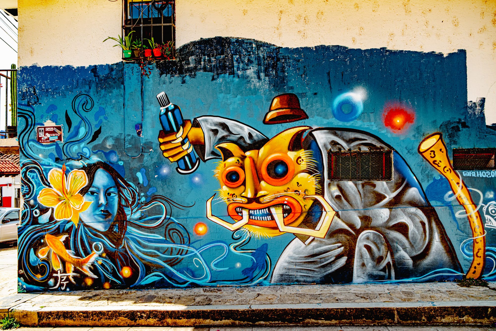 A vivid mural in El Cerrillo, San Cristóbal de las Casas, featuring a blue-toned woman with flowing hair beside a fantastical masked figure holding a spray can, set against swirling cosmic colors—blending mythology, surrealism, and contemporary Chiapas street art