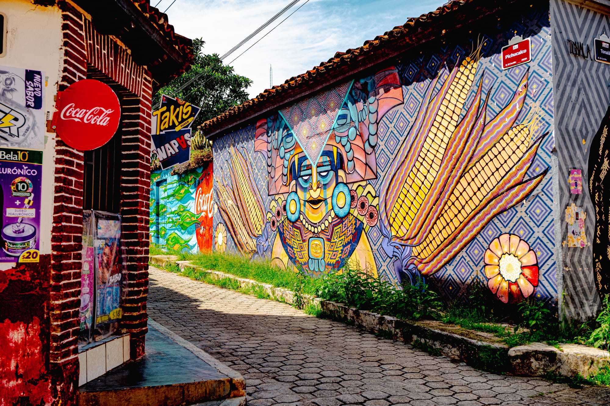 A vibrant street in El Cerrillo, San Cristóbal de las Casas, showing a large mural of a Mayan-inspired figure surrounded by maize motifs beside a small shop covered in Coca-Cola and snack ads, capturing the tension between Indigenous identity and global consumer culture in Chiapas