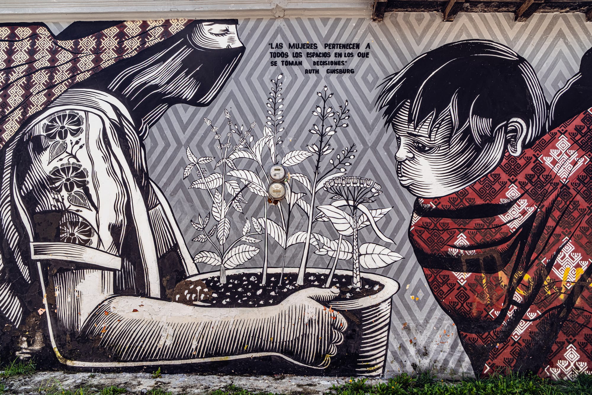 A black, white, and red mural in El Cerrillo, San Cristóbal de las Casas, depicting an Indigenous woman holding a pot of plants and a child wrapped in a patterned shawl, accompanied by a quote from Ruth Bader Ginsburg about women belonging in all spaces where decisions are made