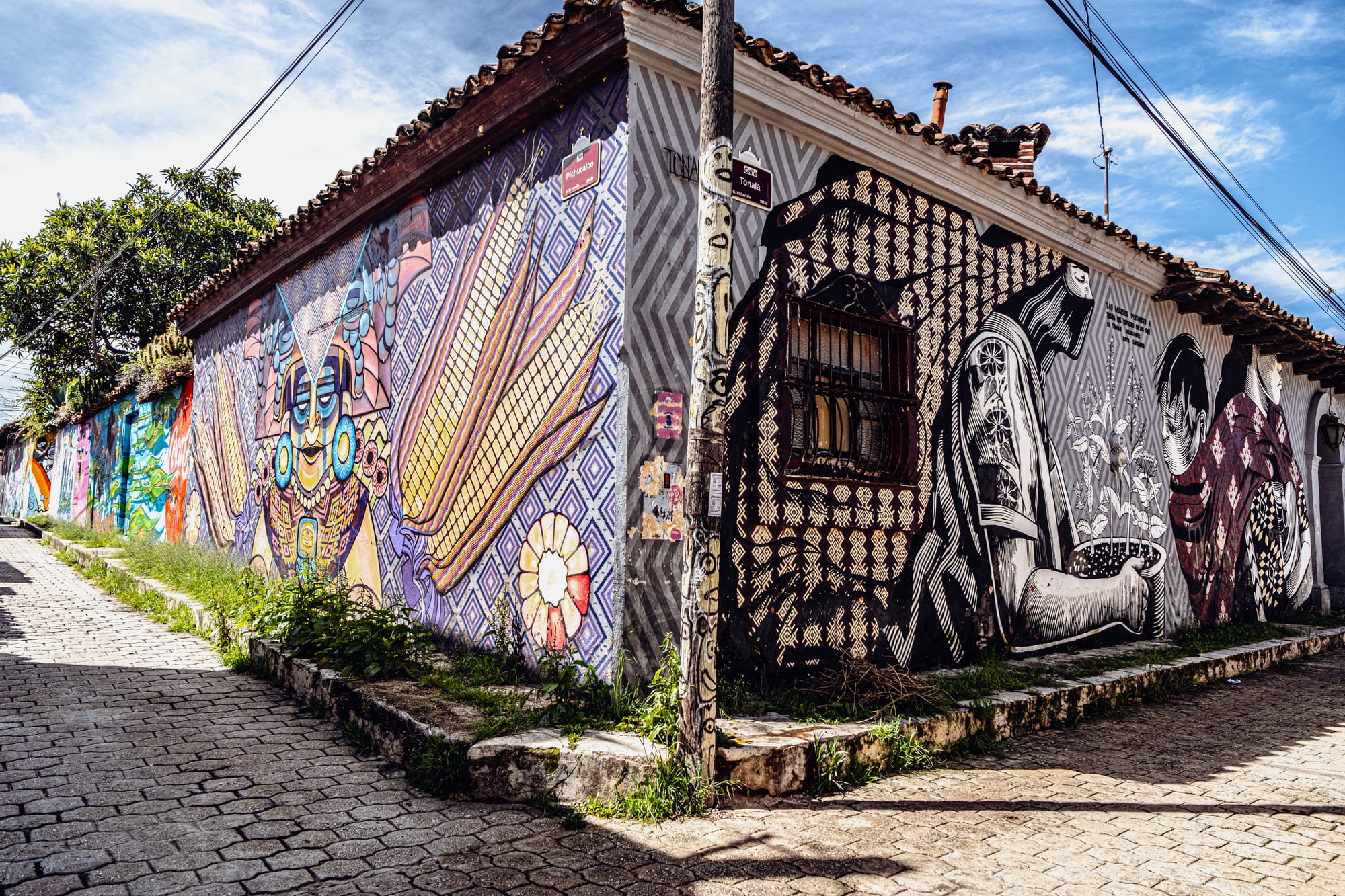 A corner building in El Cerrillo, San Cristóbal de las Casas, covered with large, detailed murals—one side showing colorful corn and Mayan-inspired patterns, the other in black and white depicting Indigenous figures sowing seeds—symbolizing Chiapas’ fusion of tradition, resistance, and contemporary street art