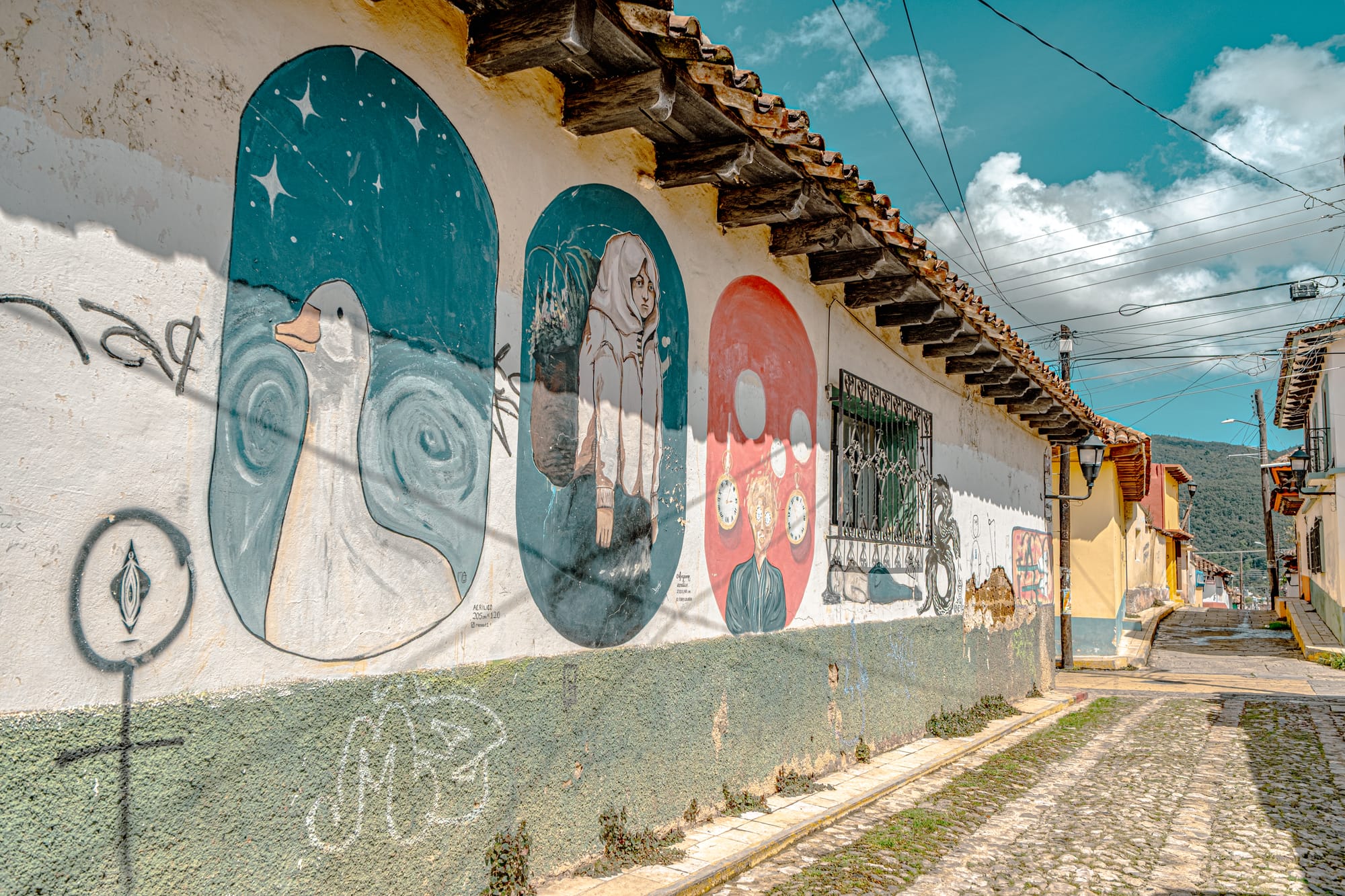 A row of murals on a sunlit street in El Cerrillo, San Cristóbal de las Casas, depicting minimalist scenes including a white goose under stars, a woman carrying plants, and a figure surrounded by clocks, blending surrealism and quiet social reflection