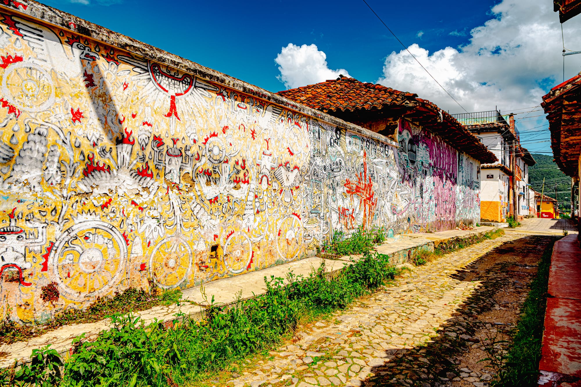 A long cobblestone alley in El Cerrillo, San Cristóbal de las Casas, lined with intricate murals featuring sun motifs, wheels, and abstract figures in yellow, red, and white tones, extending along traditional tiled-roof houses under a bright blue Chiapas sky