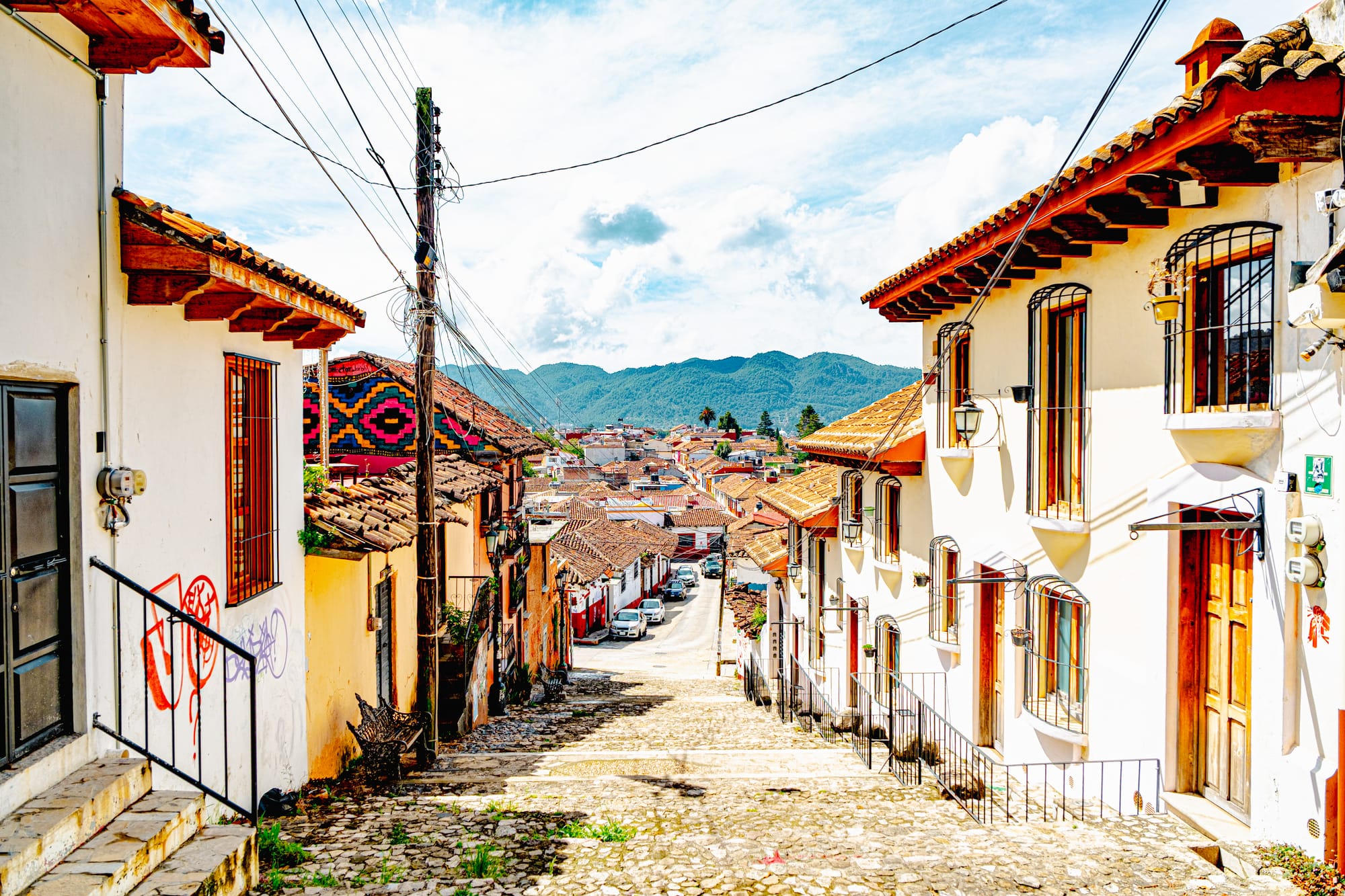 A steep cobblestone street in El Cerrillo, San Cristóbal de las Casas, lined with white adobe houses and terracotta roofs, descending toward the city and distant green mountains under a bright Chiapas sky