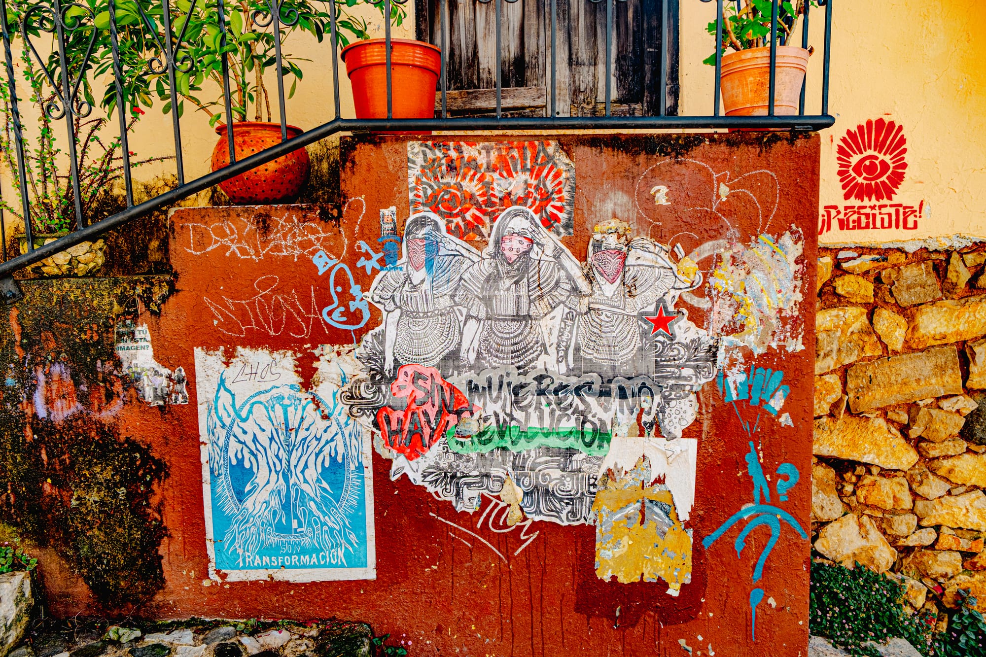 A weathered wall in El Cerrillo, San Cristóbal de las Casas, covered with feminist and Zapatista-themed street art showing three Indigenous women wearing bandanas, surrounded by graffiti and posters, with the phrase “Sin mujeres no hay revolución” (“Without women, there is no revolution”) painted across it