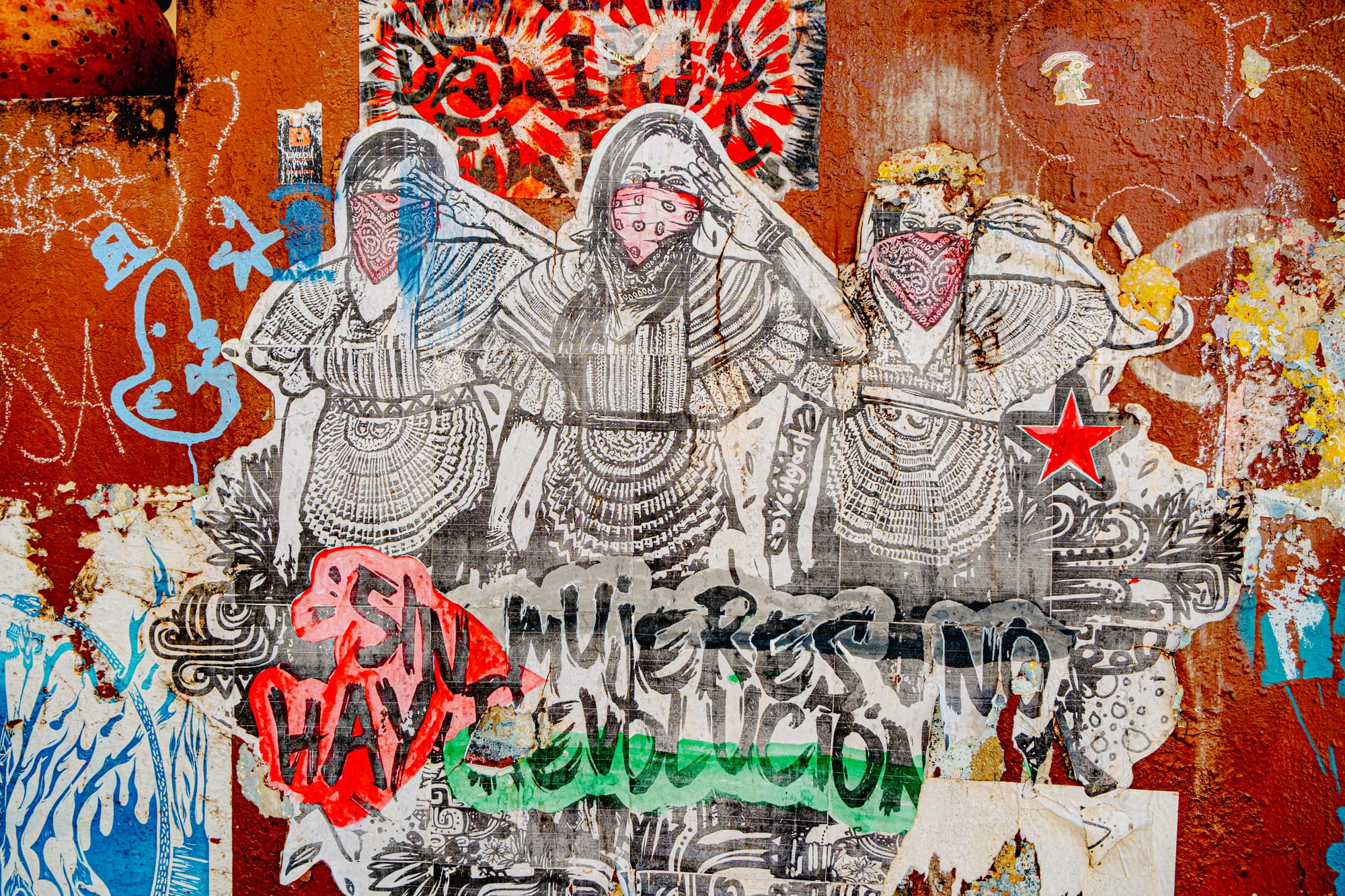A weathered stencil mural in El Cerrillo, San Cristóbal de las Casas, depicting three Indigenous women wearing traditional dresses and red bandanas, raising their fists and saluting beneath graffiti that reads “Sin mujeres no hay revolución” (“Without women, there is no revolution”), symbolizing feminist and Indigenous resistance in Chiapas