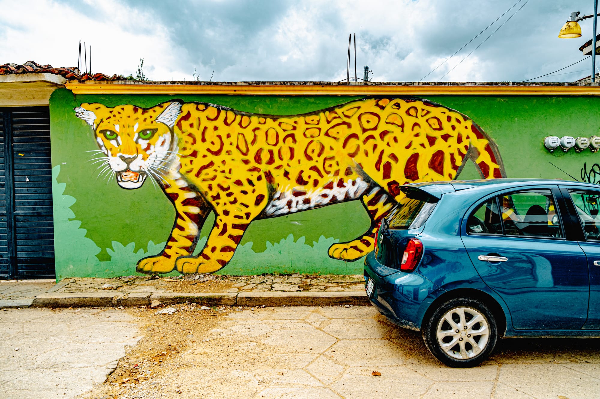 A large mural of a yellow jaguar with green eyes painted on a bright green wall in El Cerrillo, San Cristóbal de las Casas, partially obscured by a parked blue car, symbolizing power, protection, and Chiapas’ deep Indigenous heritage