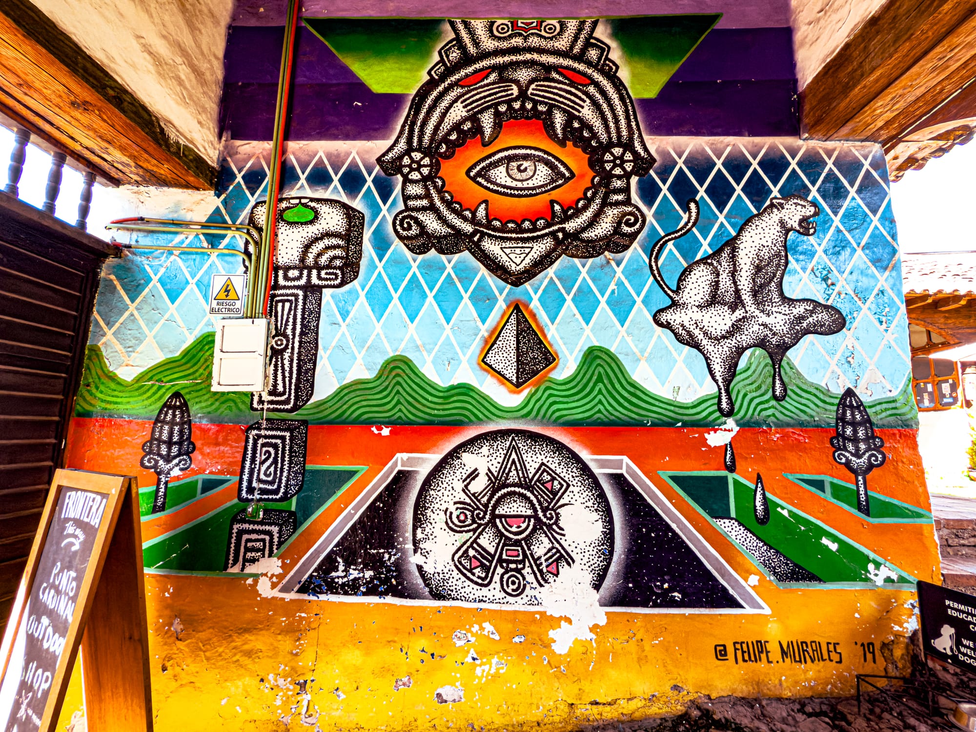 A detailed mural by artist Felipe Morales in El Cerrillo, San Cristóbal de las Casas, featuring an all-seeing eye within a jaguar-like mask, geometric mountains, and surreal black-and-white figures, merging Mayan cosmology with contemporary Mexican street art