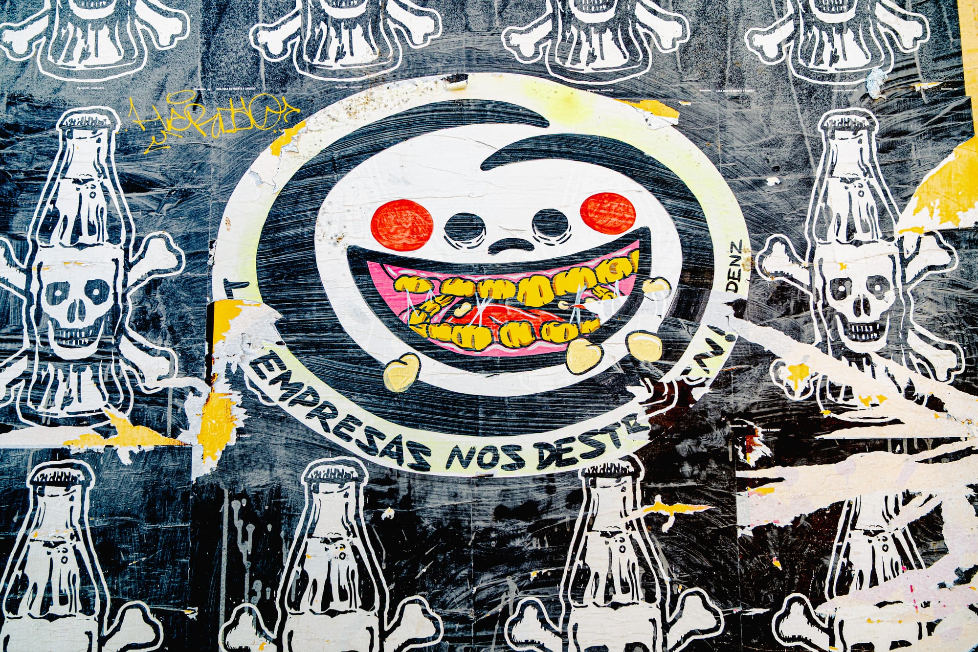 A weathered street poster in El Cerrillo, San Cristóbal de las Casas, depicting a grinning cartoon face surrounded by skulls made from soda bottles, with the phrase “Empresas nos destruyen” (“Corporations destroy us”), critiquing the environmental and social impact of corporate power in Chiapas