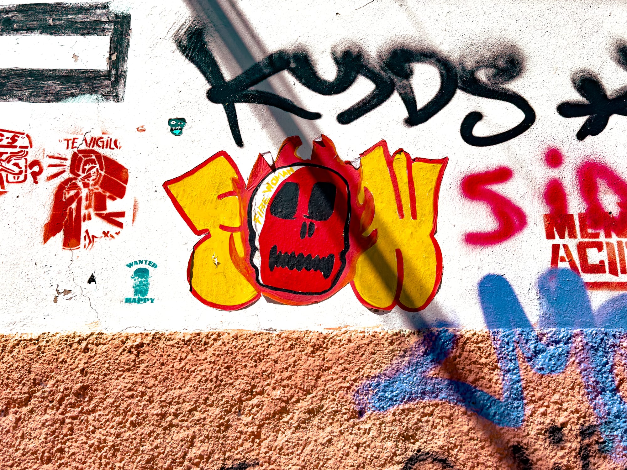 A graffiti-covered wall in El Cerrillo, San Cristóbal de las Casas, featuring a red skull with black eyes framed by yellow flames beside a red stencil of a hooded figure labeled “Te Vigilo,” combining local street art with themes of rebellion and surveillance