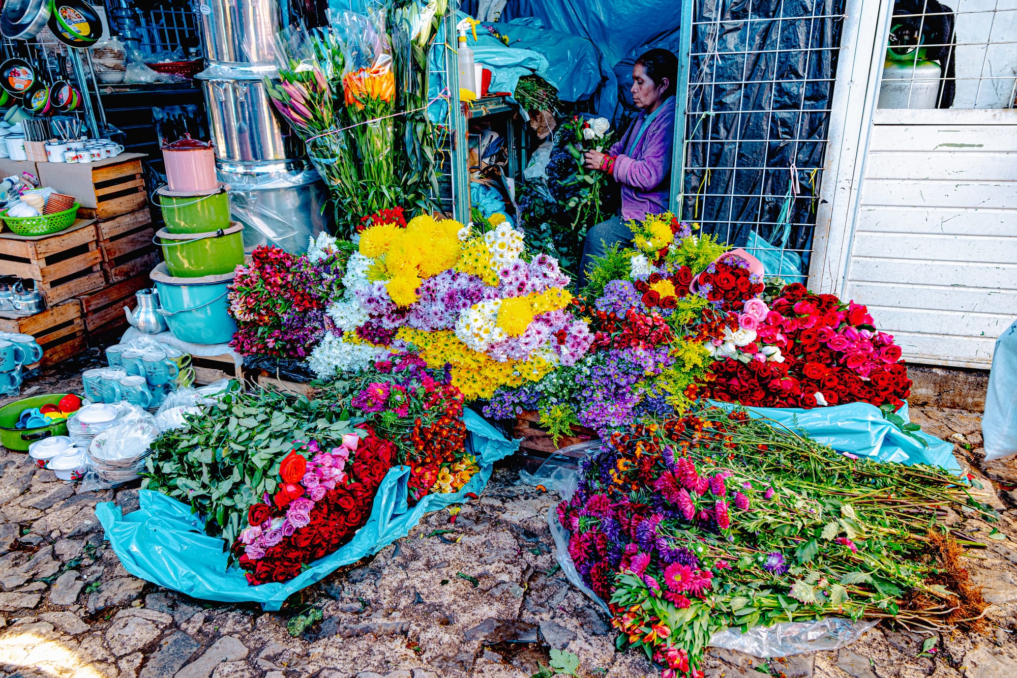 Vendor arranging colorful flowers, including roses and marigolds, at Santo Domingo Market in San Cristóbal de las Casas, Chiapas, surrounded by enamel cookware and cobblestone streets