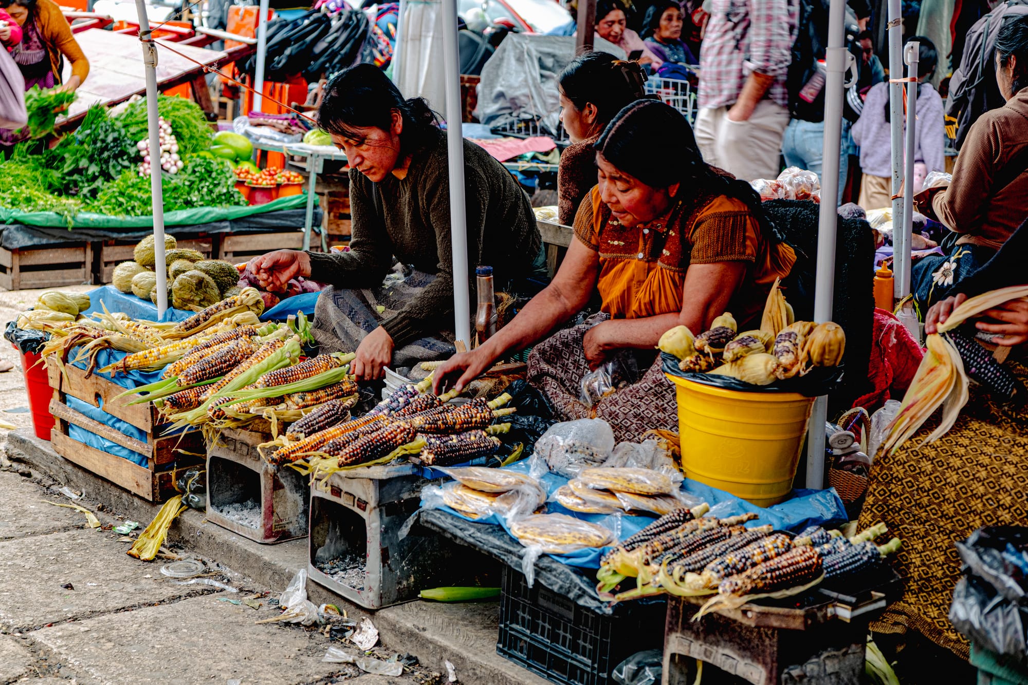 Two women roasting and selling colorful native corn at Santo Domingo Market in San Cristóbal de las Casas, Chiapas, surrounded by crates of maize and local produce