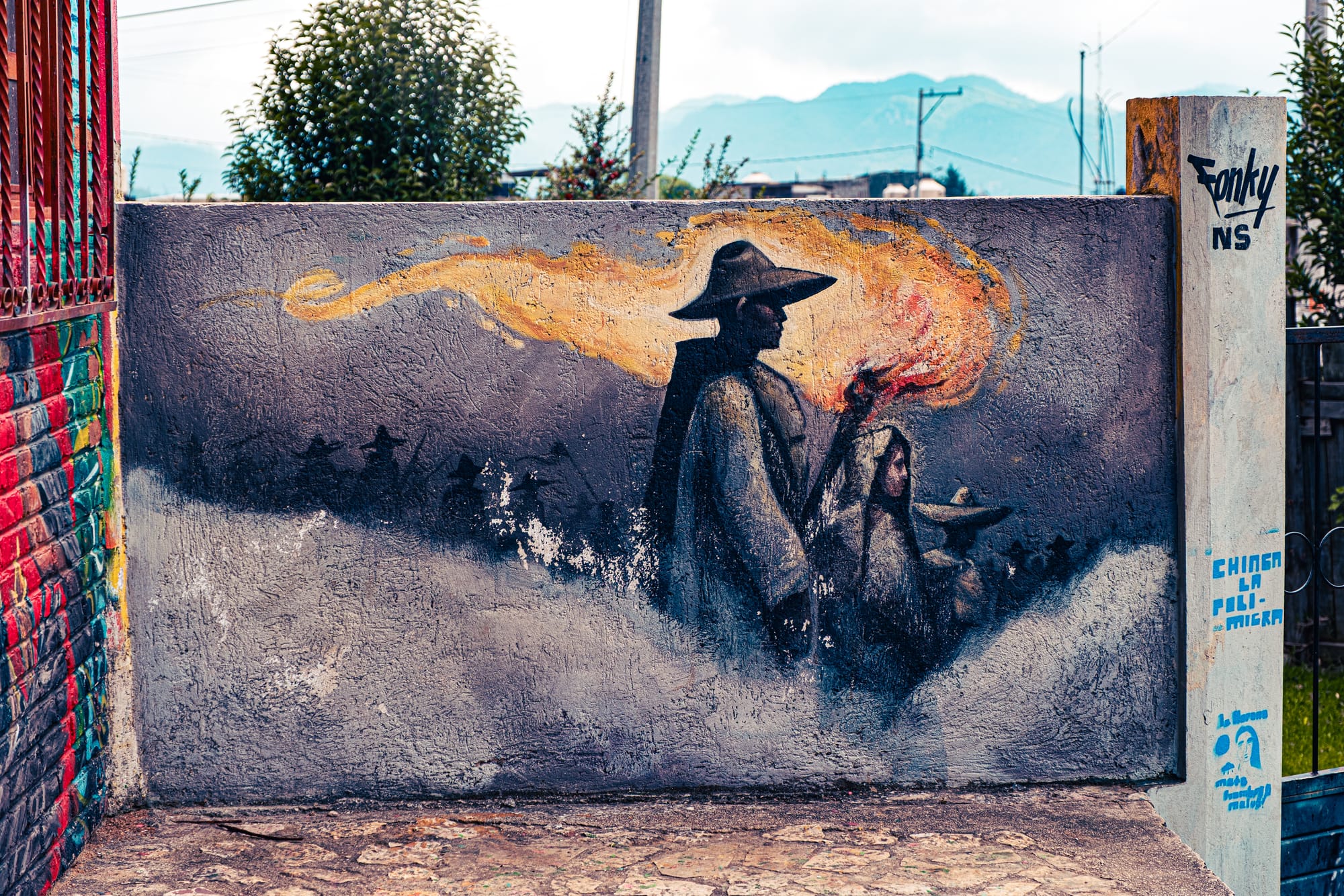 Mural at CIDECI showing shadowed figures in wide-brimmed hats emerging from mist, led by two people holding a flaming torch, painted on a textured wall with mountains faintly visible in the background