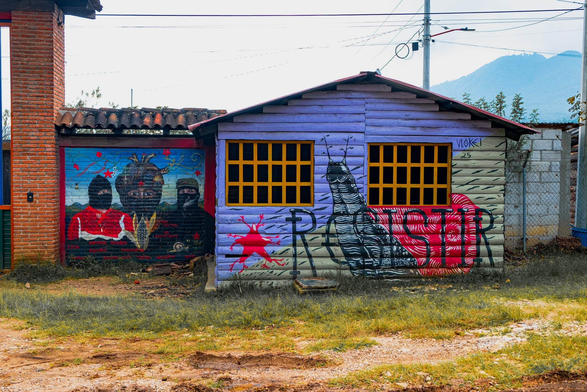 Two adjacent buildings at CIDECI painted with murals—one showing masked Zapatista figures with stars and maize, and the other featuring a large black-and-white insect-like figure and red lettering—set against a rural backdrop with mountains and power lines