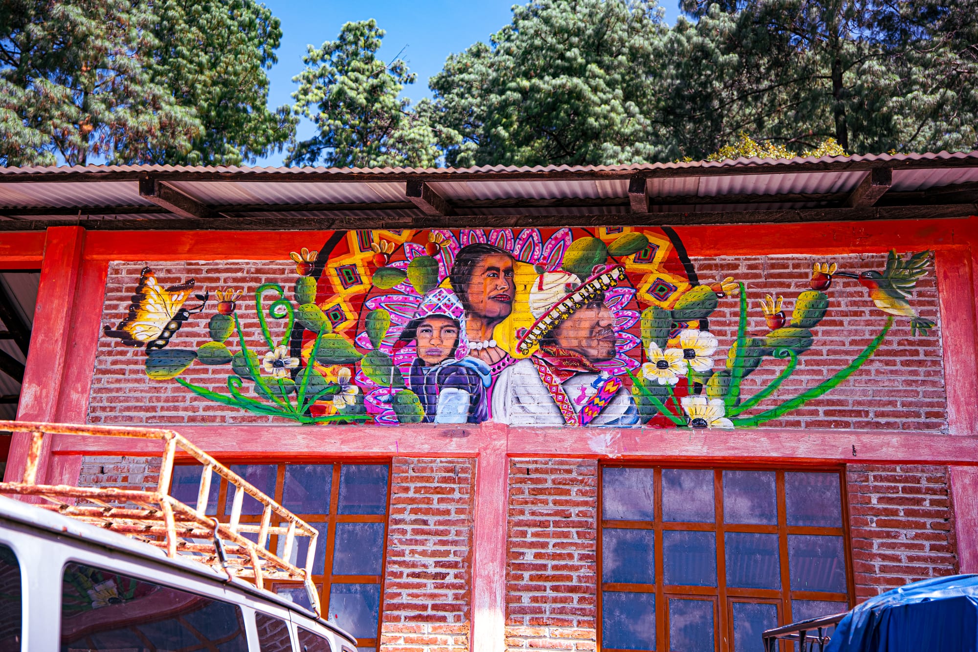 Mural at CIDECI depicting three Indigenous figures surrounded by colorful flowers, cactus pads, butterflies, and a hummingbird, painted on a brick building beneath a metal roof with tall pine trees in the background