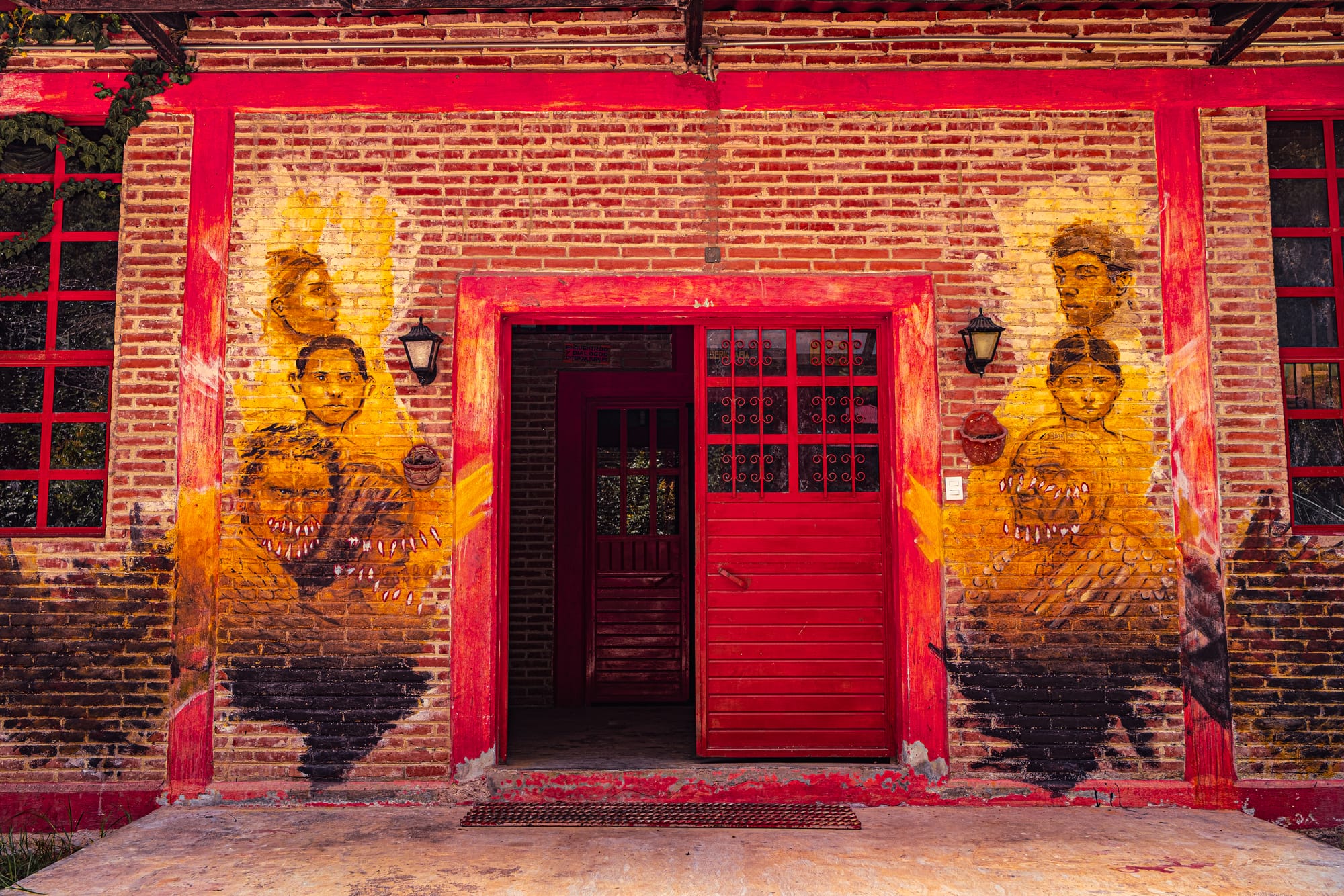 Brick building at CIDECI with a red doorway and symmetrical murals on each side, depicting layered portrait-style faces painted in warm yellow and brown tones, creating a textured, expressive entrance
