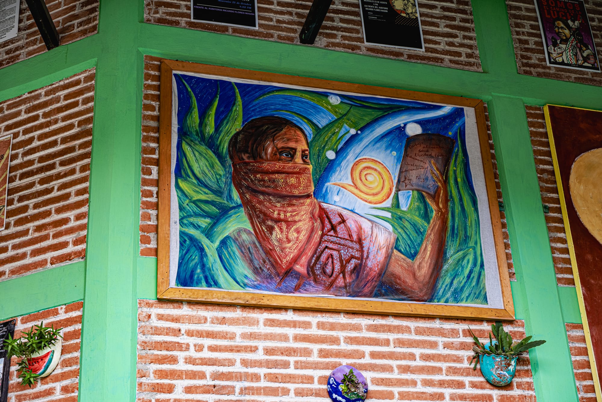 Painting at CIDECI depicting a masked figure holding a book against a swirling cosmic background with plants and bright colors, framed on a brick wall with green trim