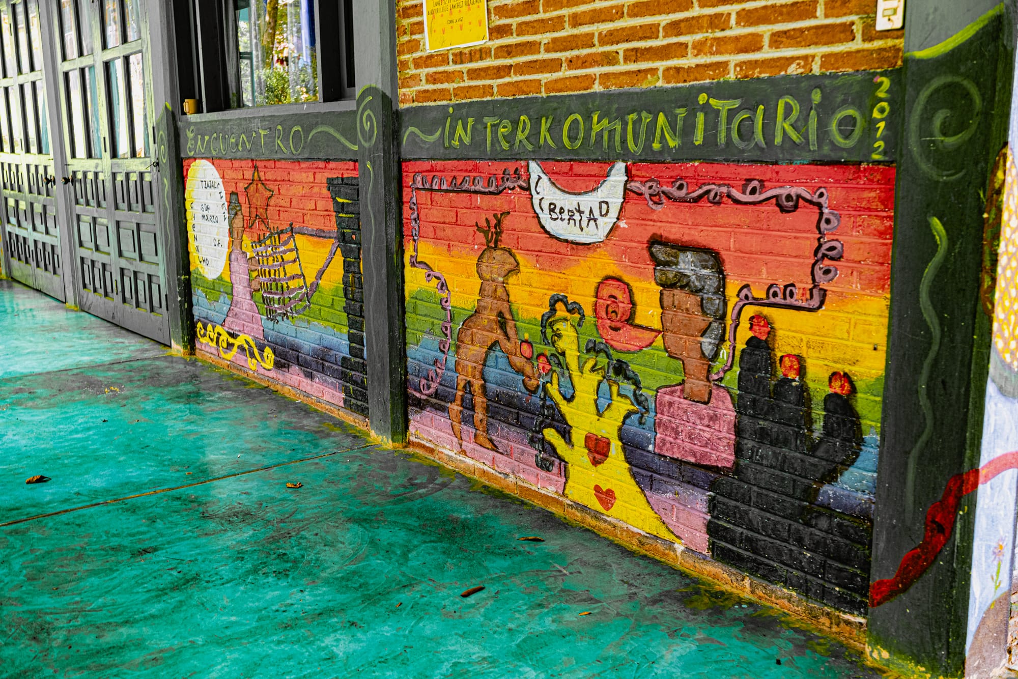 Small fenced building at CIDECI painted with a vibrant mural of snails, ladybugs, plants, and abstract nature motifs, set against a bright sky and surrounding greenery