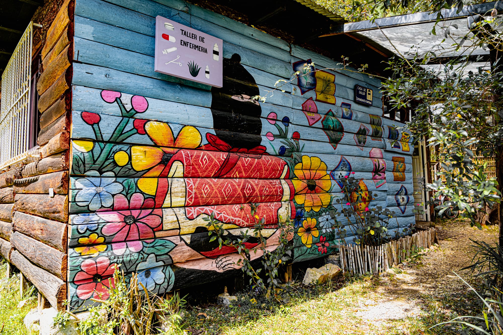 Mural-covered exterior of the infirmary at CIDECI-UniTierra, showing a masked figure in traditional clothing surrounded by bright flowers and colorful painted motifs on a wooden building