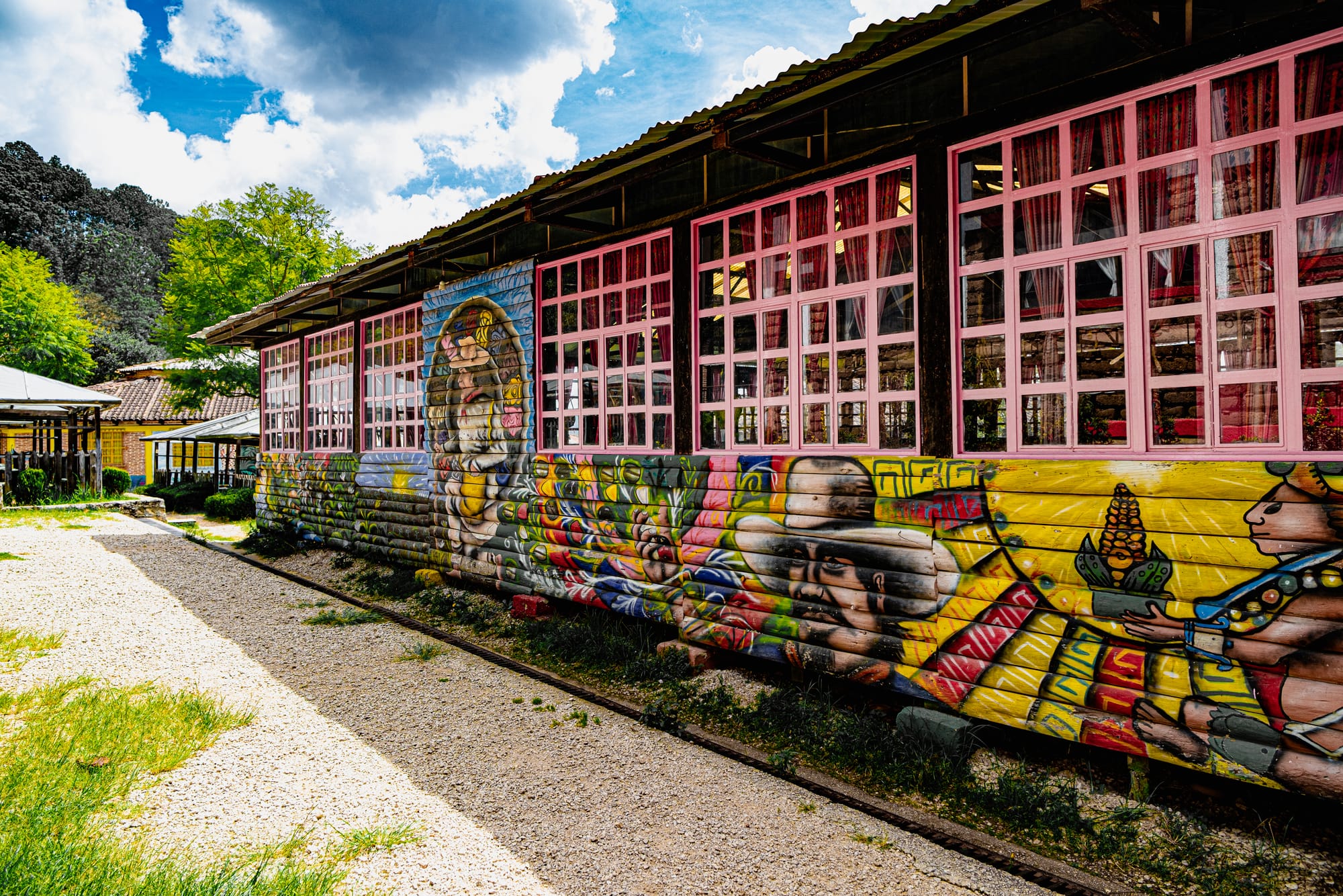Long wooden building at CIDECI with pink-framed windows and a vibrant mural depicting Indigenous figures, corn, flowers, and cultural motifs along the exterior wall, set against trees and a bright sky
