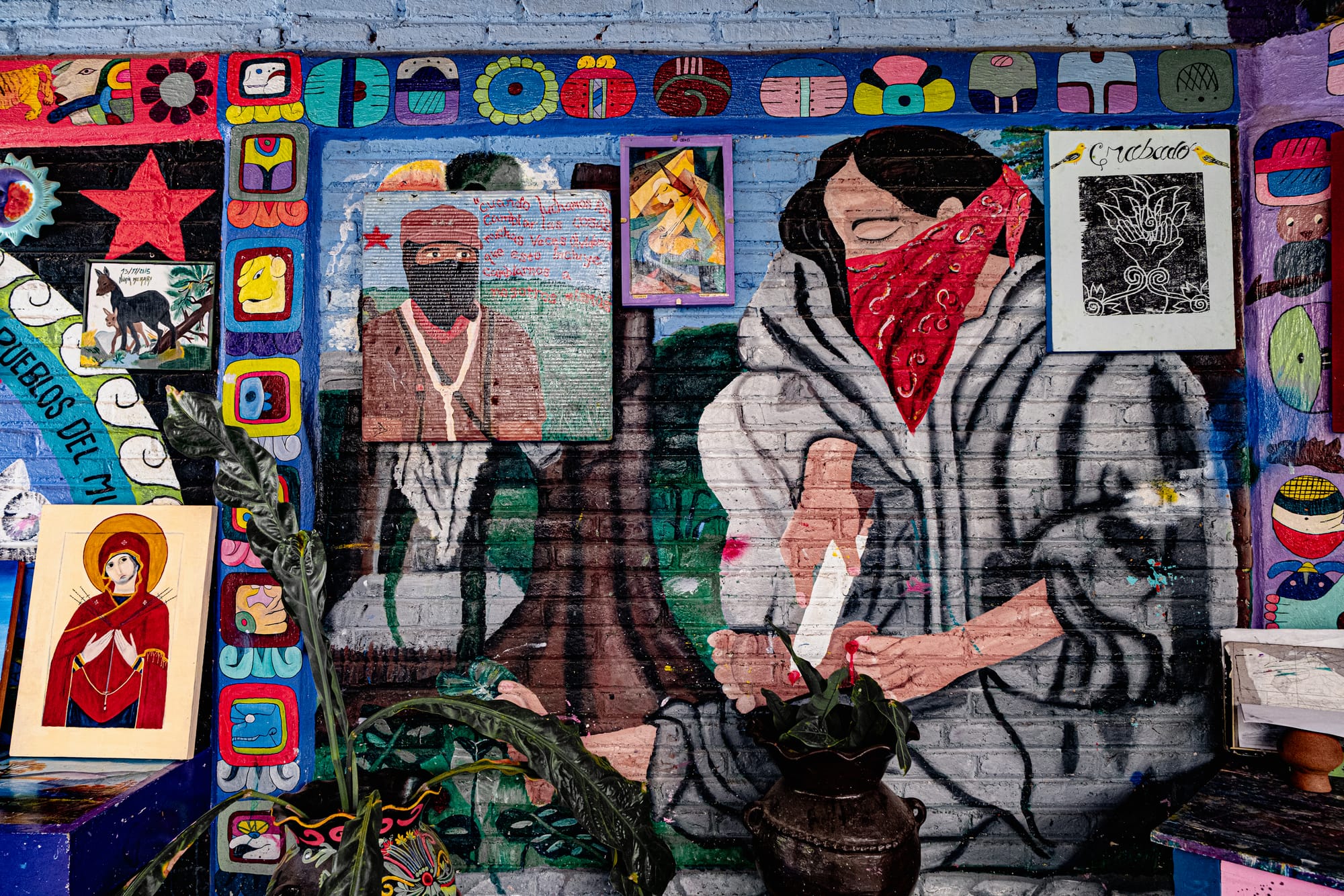 Mural at CIDECI depicting a masked woman in traditional clothing, alongside a painted portrait of a Zapatista figure, surrounded by colorful Mayan-inspired borders, symbols, and additional artwork displayed on the wall.