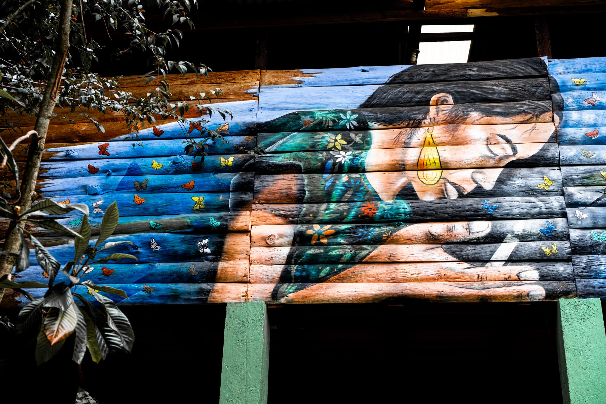 Mural at CIDECI depicting an Indigenous woman in a floral blouse leaning forward to cup a small light in her hands, surrounded by colorful butterflies against a blue sky background on wooden panels