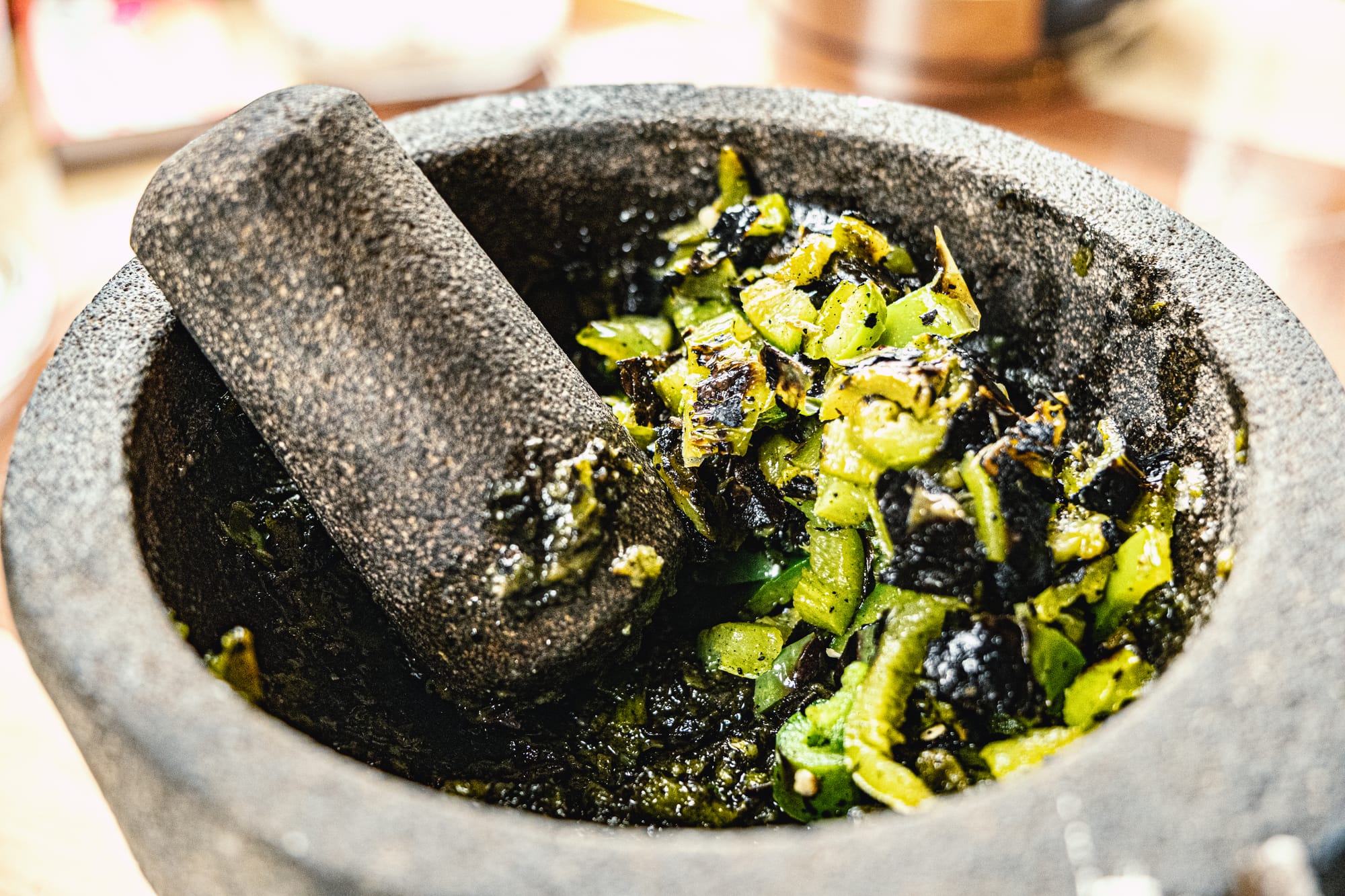 Close-up of chopped, fire-roasted jalapeños being crushed in a stone molcajete, the mixture glossy and charred