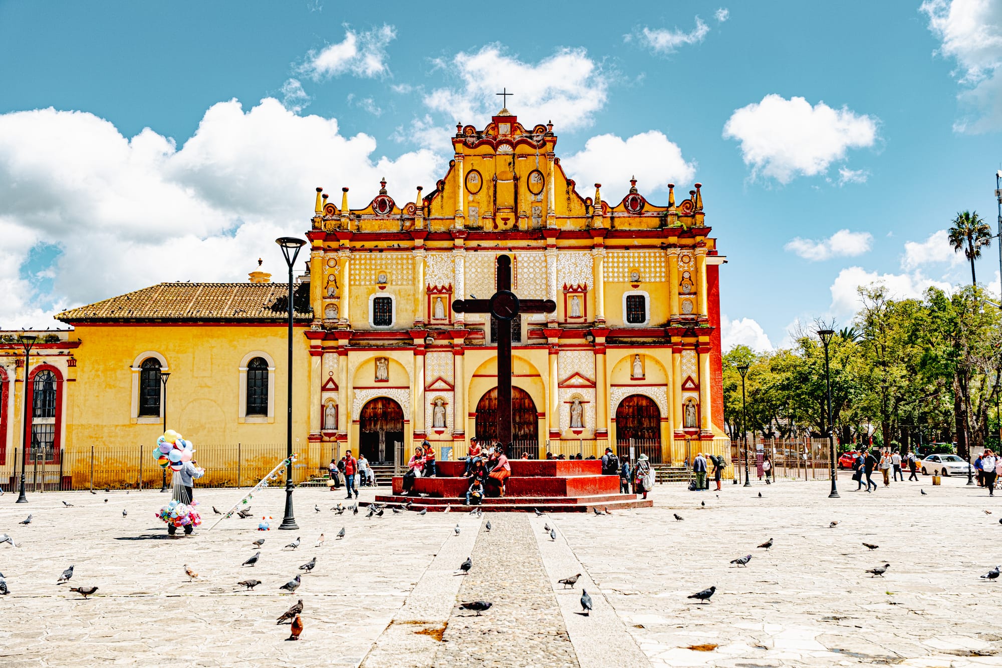 Catedral de San Cristóbal Mártir in San Cristóbal de las Casas, Chiapas, Mexico, showing the yellow Baroque cathedral façade, central cross, and Plaza 31 de Marzo with people and pigeons