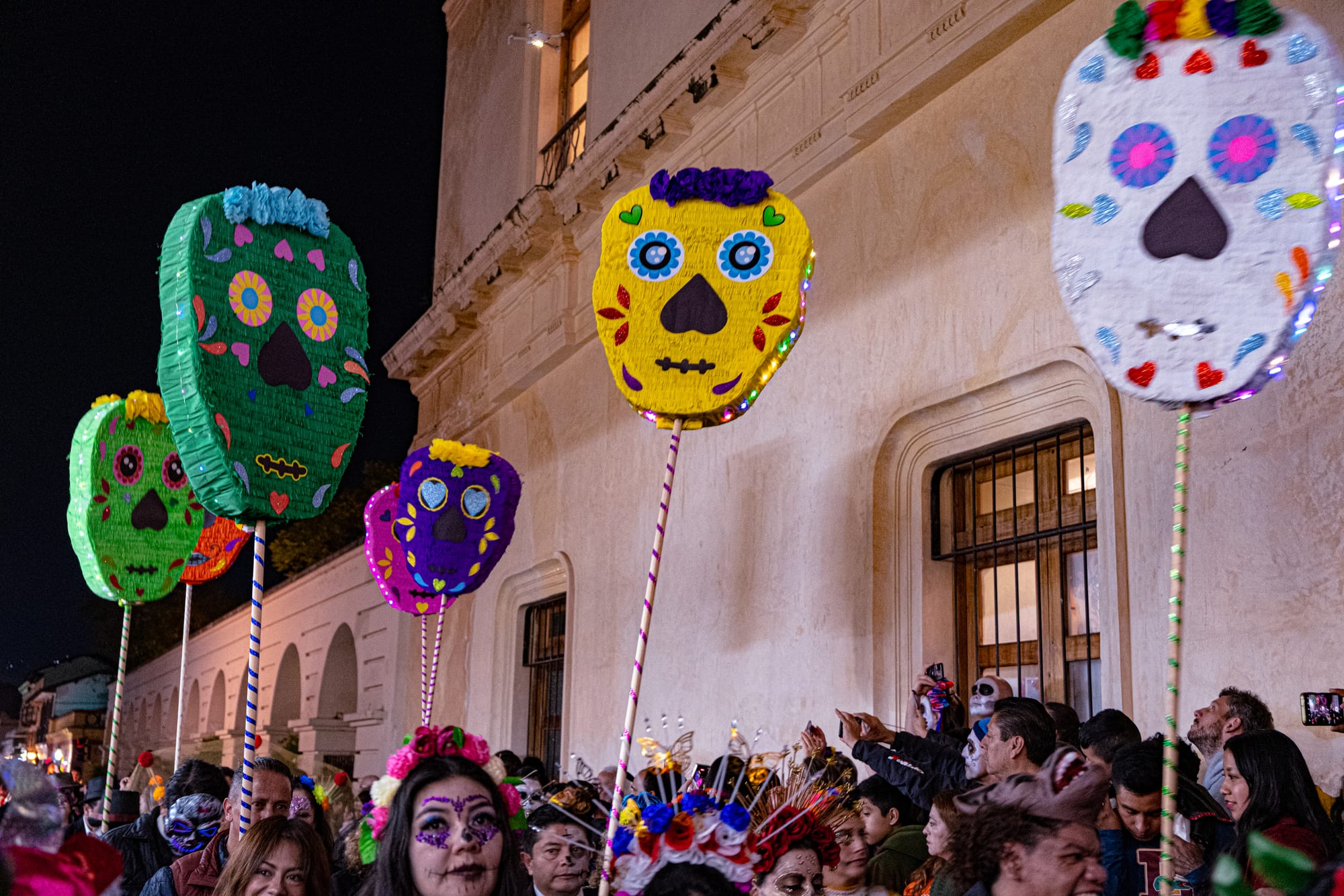 Day of the Dead parade in San Cristóbal de las Casas, Chiapas, Mexico, large colorful skull lanterns carried above the crowd, people wearing face paint and floral headpieces, nighttime Día de Muertos procession with illuminated calavera decorations and dense crowds in the historic center