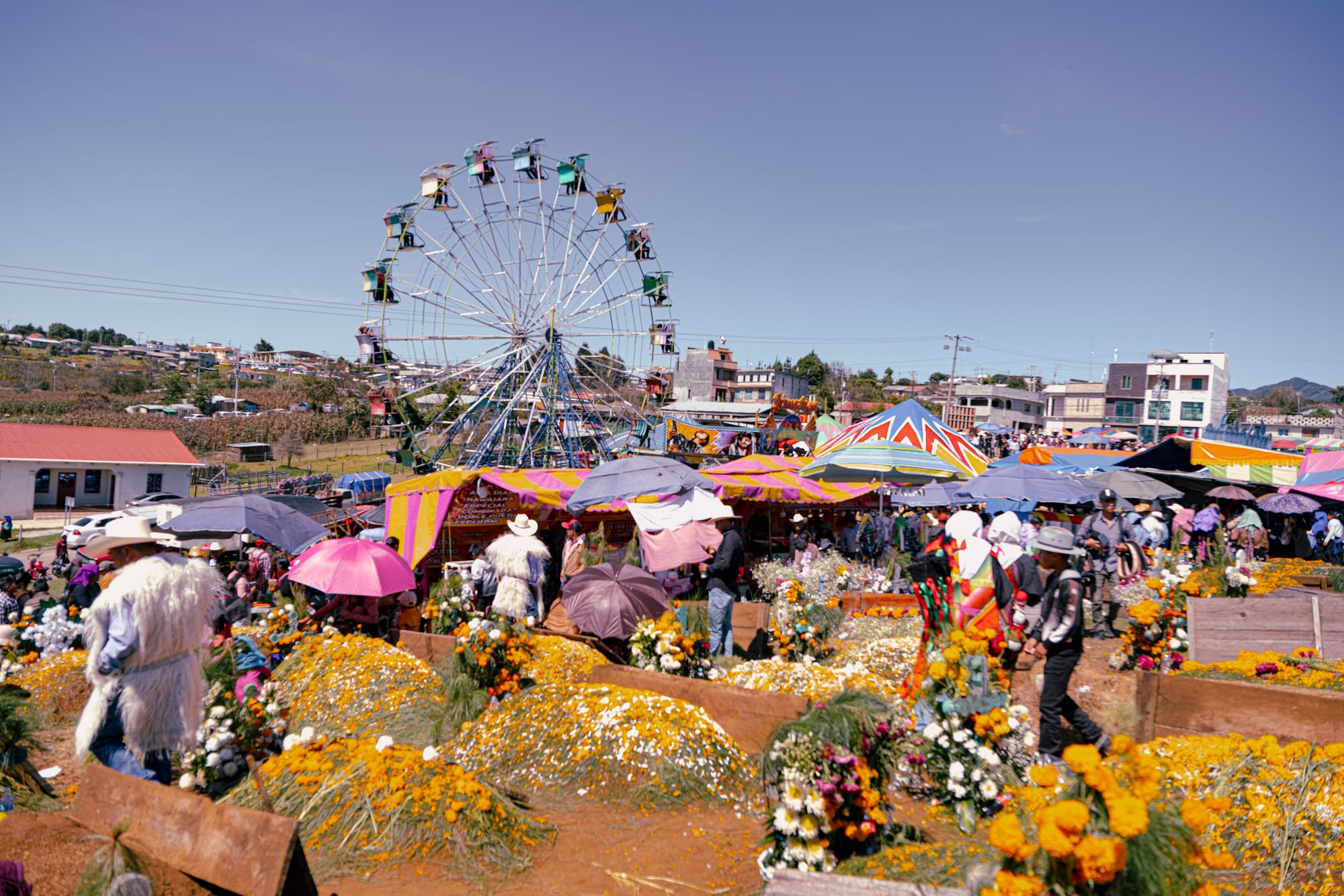 Día de Muertos at Romerillo Cemetery in San Cristóbal de las Casas, Chiapas, Mexico, decorated graves covered in marigolds and flowers, families gathering among tombs beside an amusement park with ferris wheel, local Day of the Dead cemetery celebration with strong community atmosphere