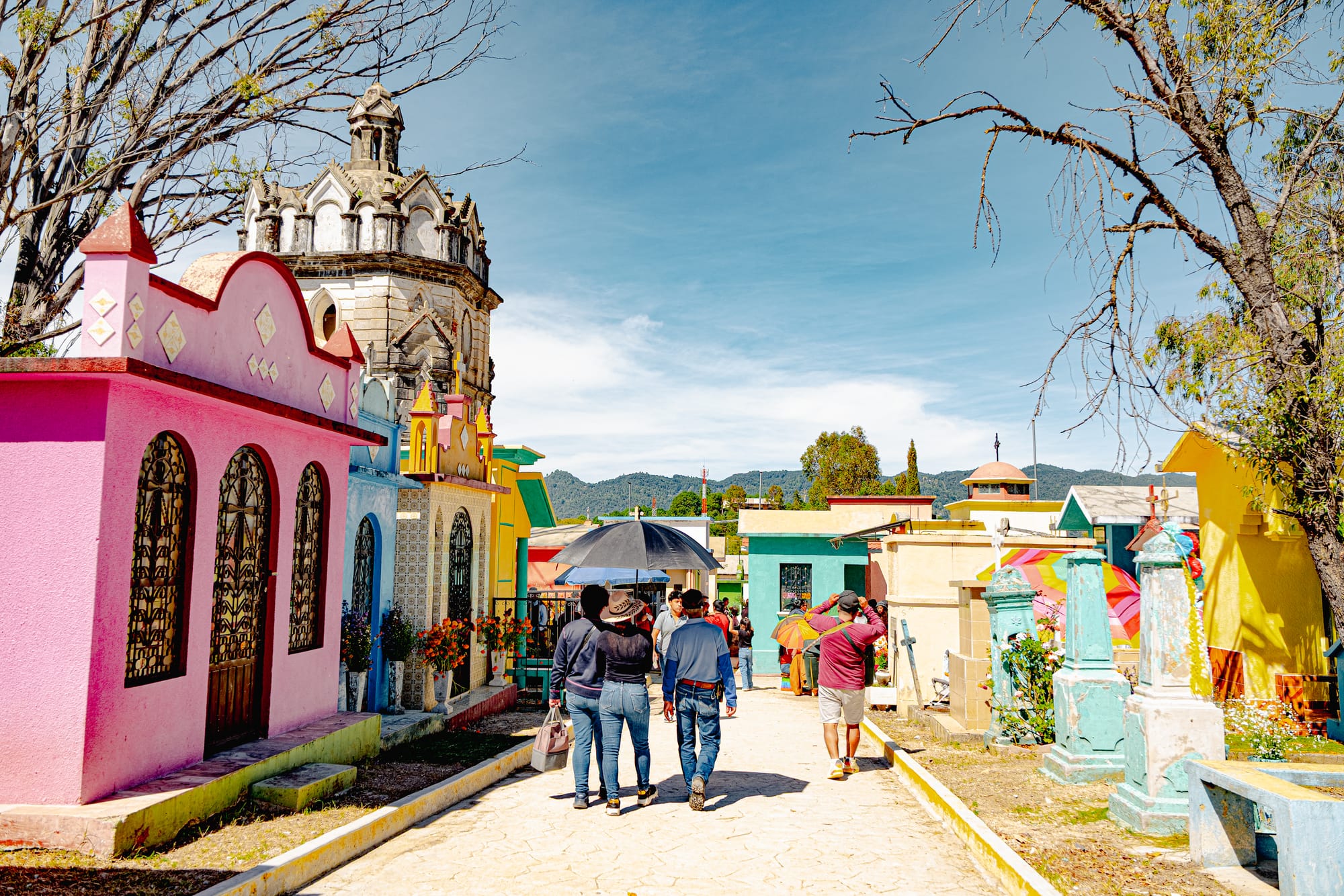 Día de Muertos at San Cristóbal Municipal Pantheon in San Cristóbal de las Casas, Chiapas, Mexico, colorful above ground tombs painted like small houses, people walking through cemetery pathways carrying flowers, traditional Day of the Dead celebrations inside the city cemetery with mountain backdrop