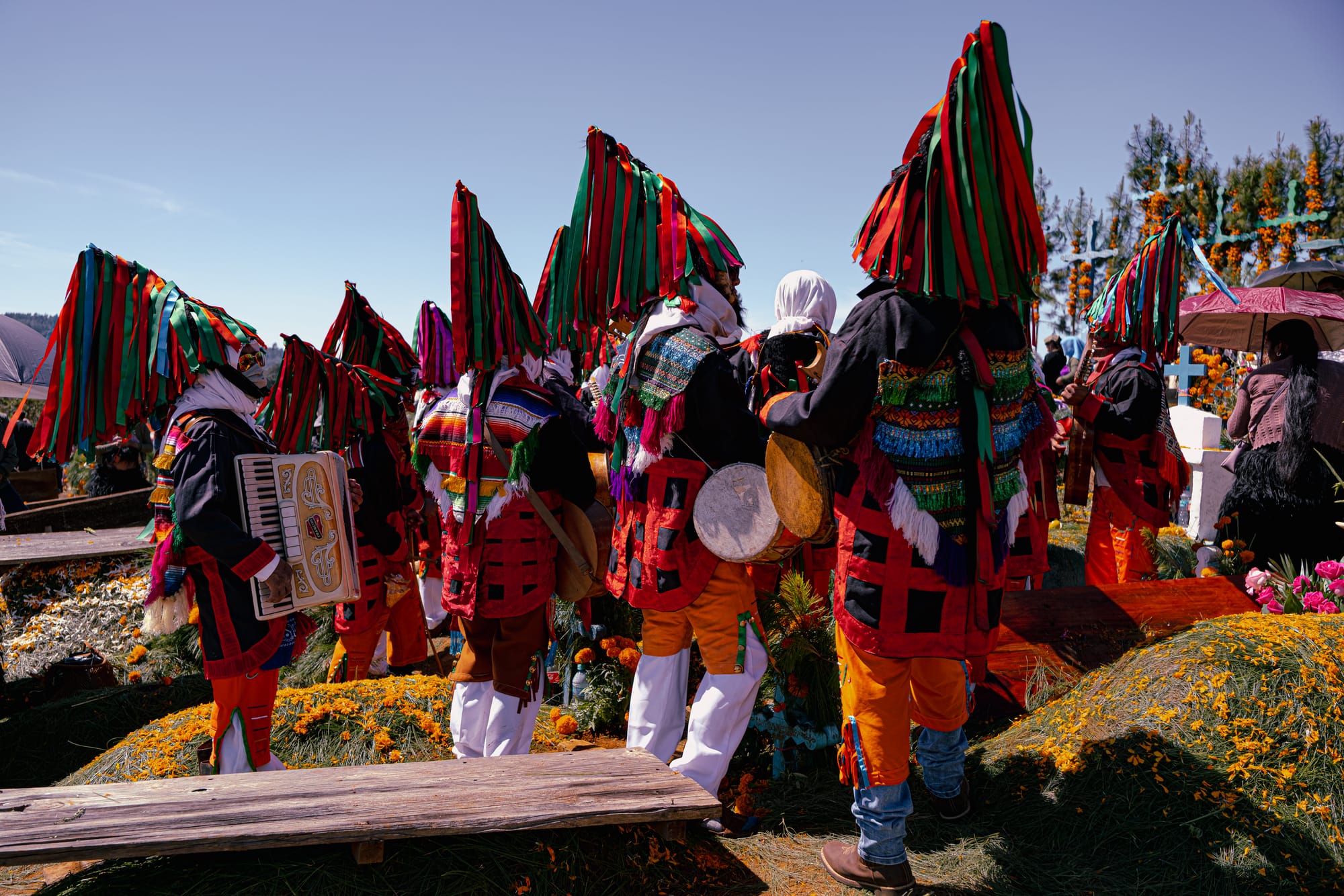 Día de Muertos at Romerillo Cemetery Chiapas, indigenous musicians wearing traditional ribbon headdresses and colorful woven clothing gathered among pine covered graves, live band with accordion drums and guitars playing during Day of the Dead celebration, marigold covered hilltop cemetery with wooden grave markers near San Cristóbal de las Casas