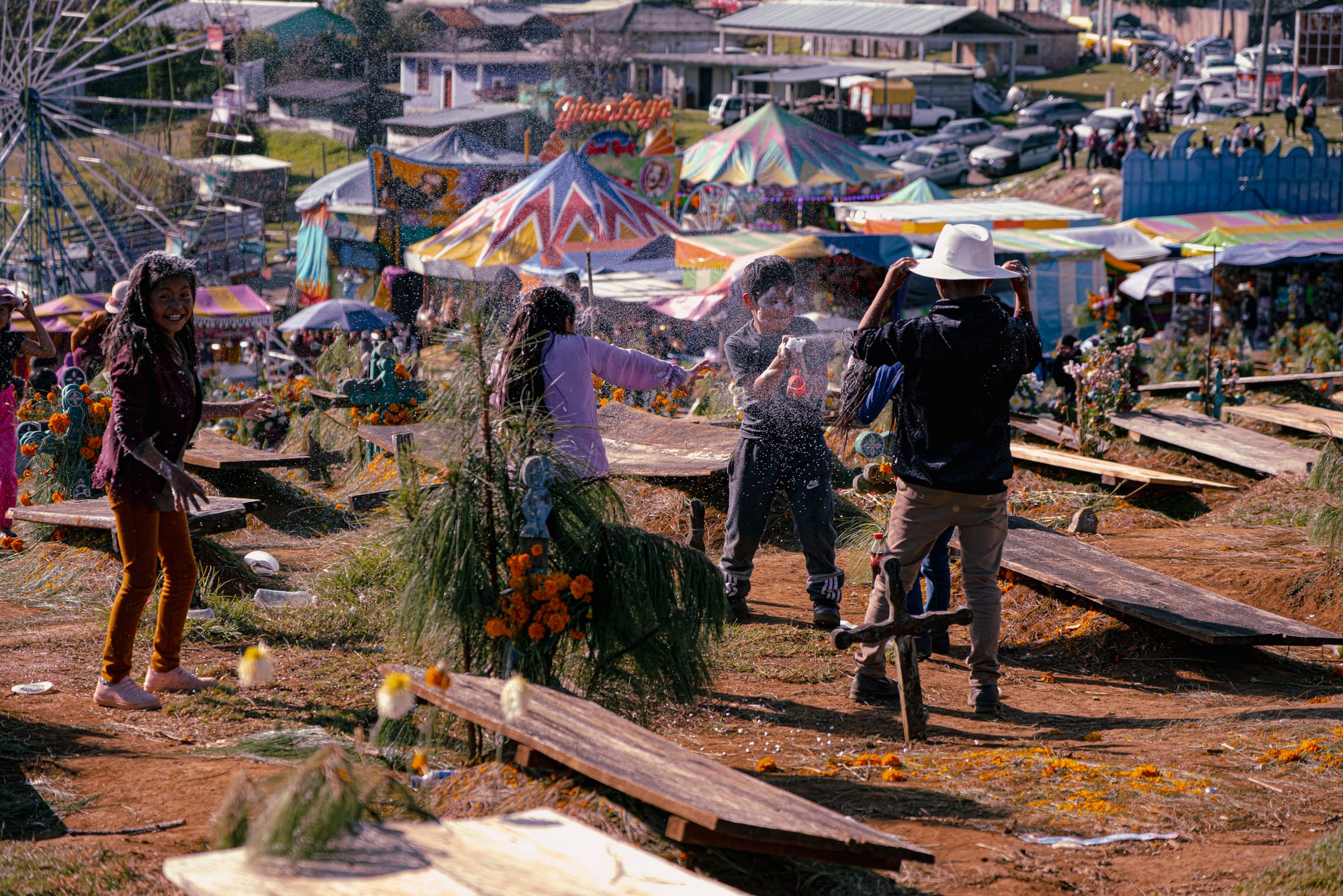 Día de Muertos at Romerillo Cemetery Chiapas, children playing and laughing among pine covered graves and marigold decorated tombs, amusement park rides visible beside the hilltop cemetery, local families celebrating Day of the Dead with joy music and community gathering near San Cristóbal de las Casas