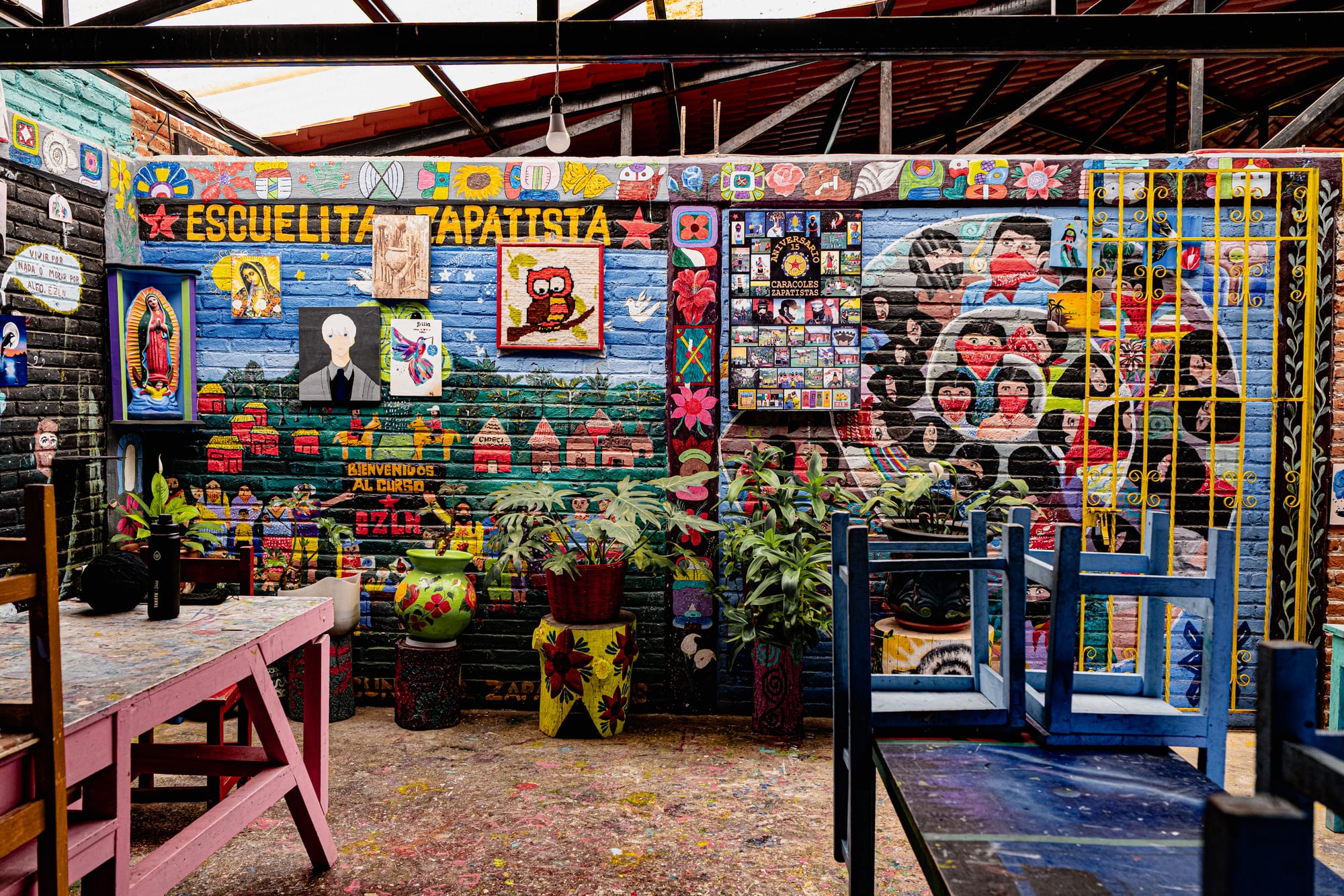 CIDECI–UniTierra courtyard in San Cristóbal de las Casas, with walls covered in colorful murals referencing indigenous autonomy and Zapatista education, surrounded by painted chairs, tables, potted plants, and a paint-splattered floor beneath an open roof