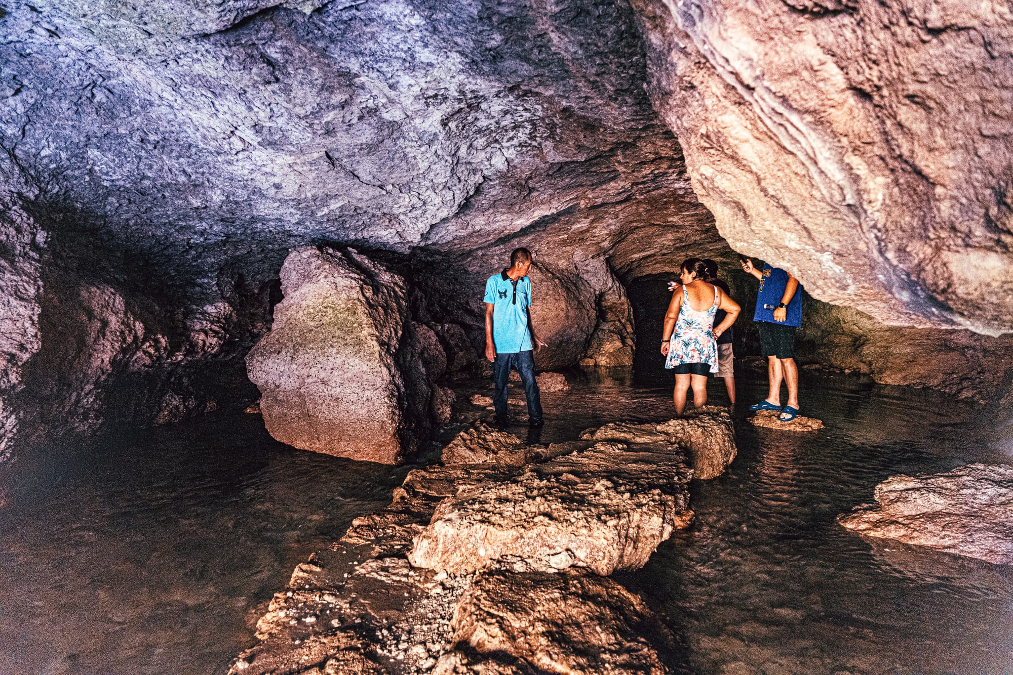 Visitors standing inside the cave behind Misol-Há waterfall near Palenque, Chiapas, wading through shallow water beneath a rocky overhang, showing the interior cave area accessible by walking behind the waterfall and one of the most unique features of visiting Misol-Há waterfall in southern Mexico