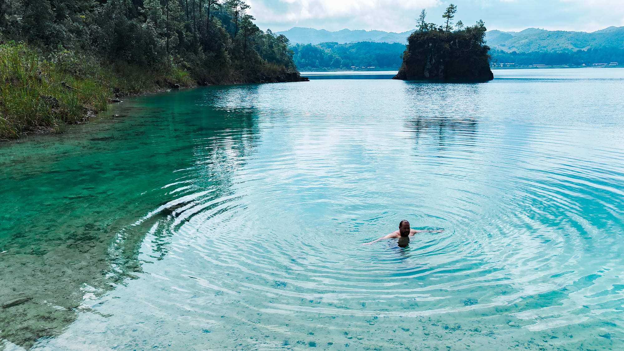 Person swimming in the clear turquoise water of Montebello Lakes in Chiapas, with forested hills, rocky outcrops, and calm ripples spreading across the lake