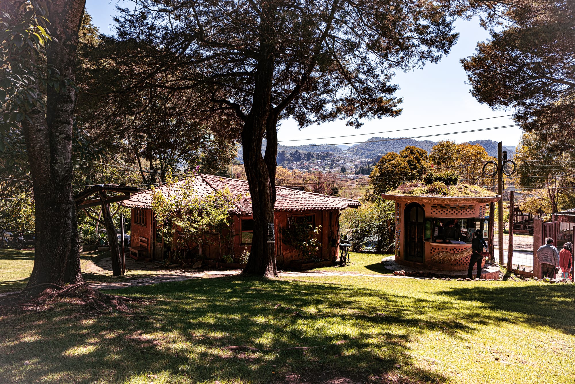 Entrance area of Moxviquil in San Cristóbal de las Casas, with an earthen-roofed ticket booth, shaded trees, and views of the surrounding highland hills in the background