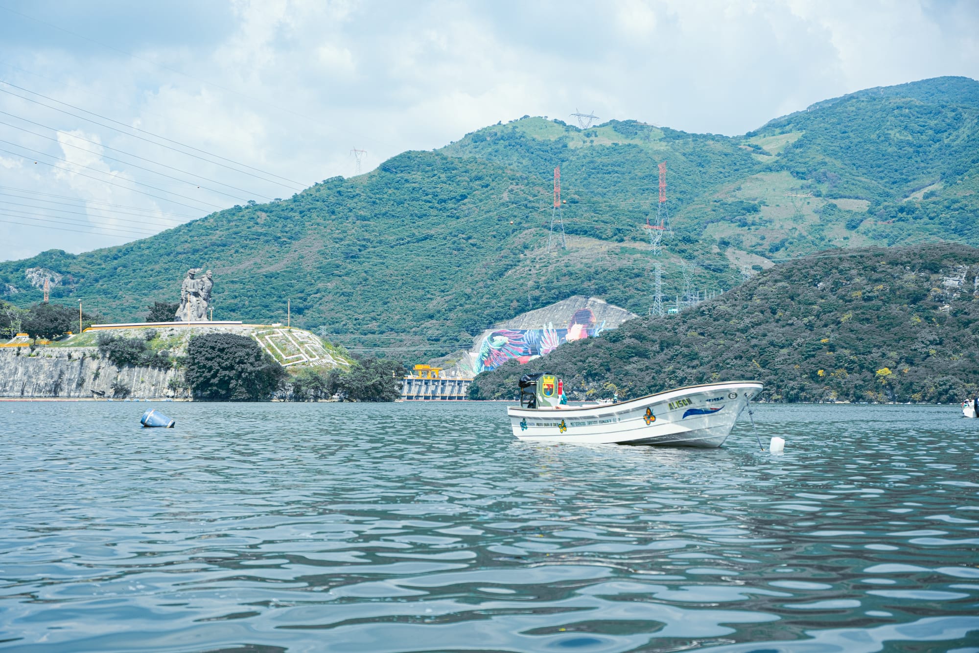 Boat floating on the Grijalva River near the entrance of Sumidero Canyon, with the monument at the Chicoasén Dam and surrounding green hills visible in the background