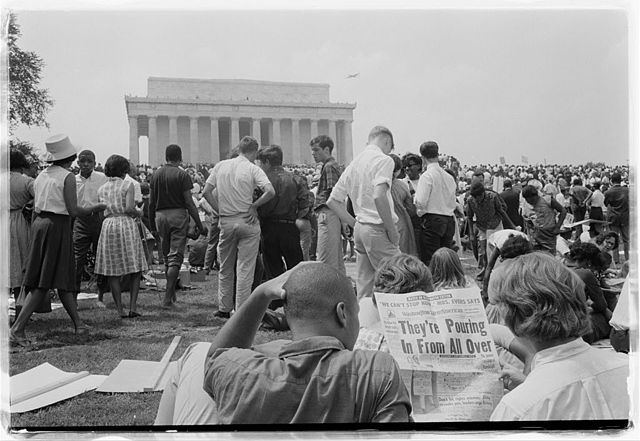Photograph shows a crowd of African Americans and whites on the grounds of the Lincoln Memorial; two men in foreground read a newspaper with the headline: "They're Pouring In From All Over."
