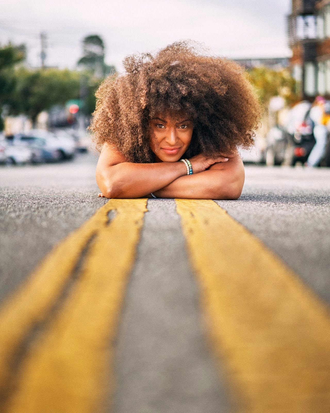 A woman with tight, voluminous curls lying on her stomach on the double yellow stripes on the middle of a road.