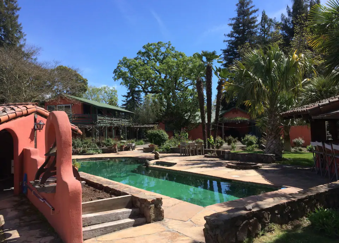 A large pool with a small stucco structure with an arch in the foreground, and a large house in the far background.