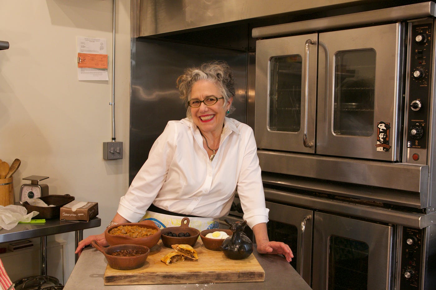 Paula Tejeda smiling in an industrial kitchen, standing over bowls of ingredients on a butcher block.