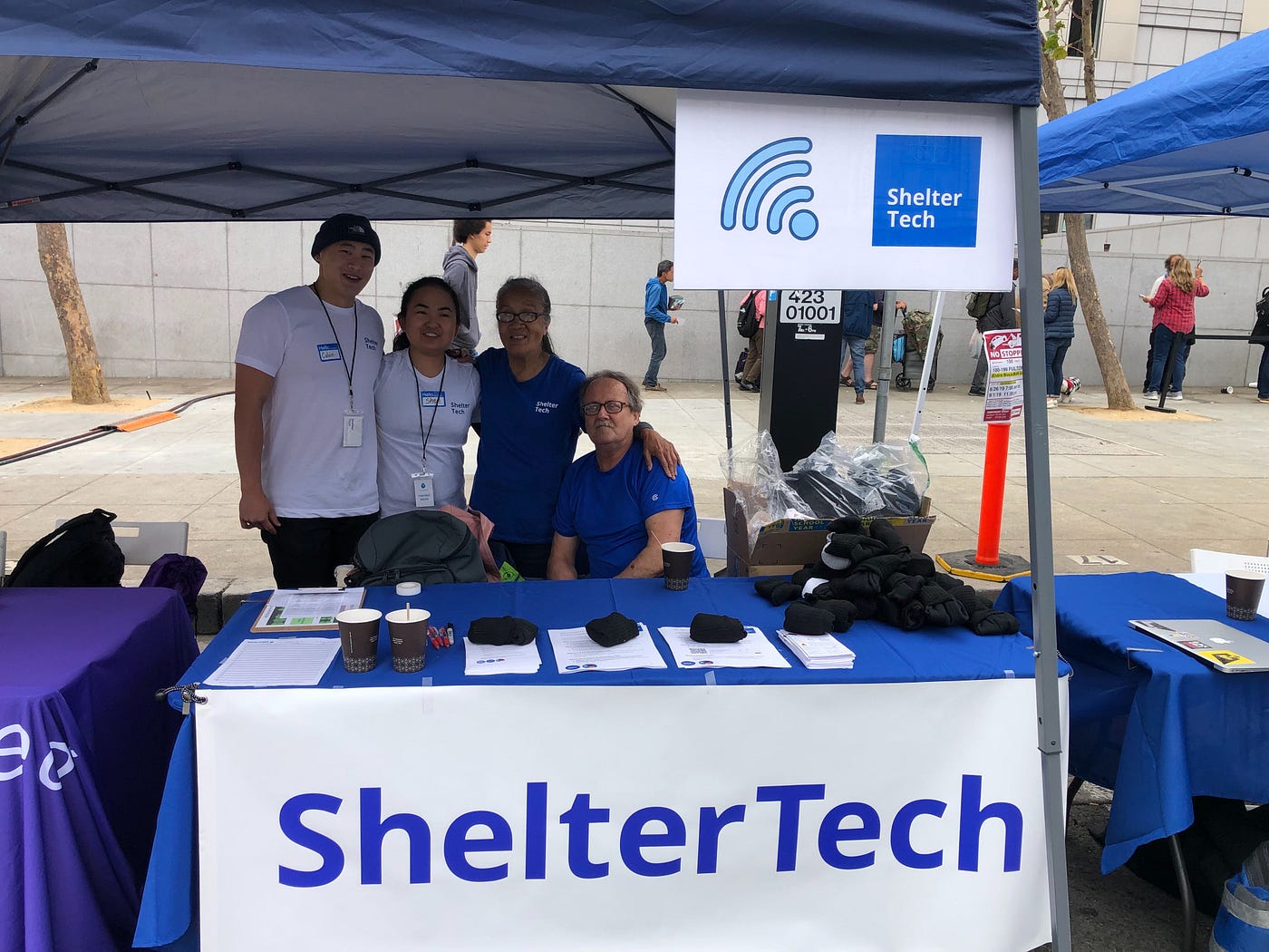 4 people wearing lanyards standing behind a table under a tent with a banner that says “ShelterTech”