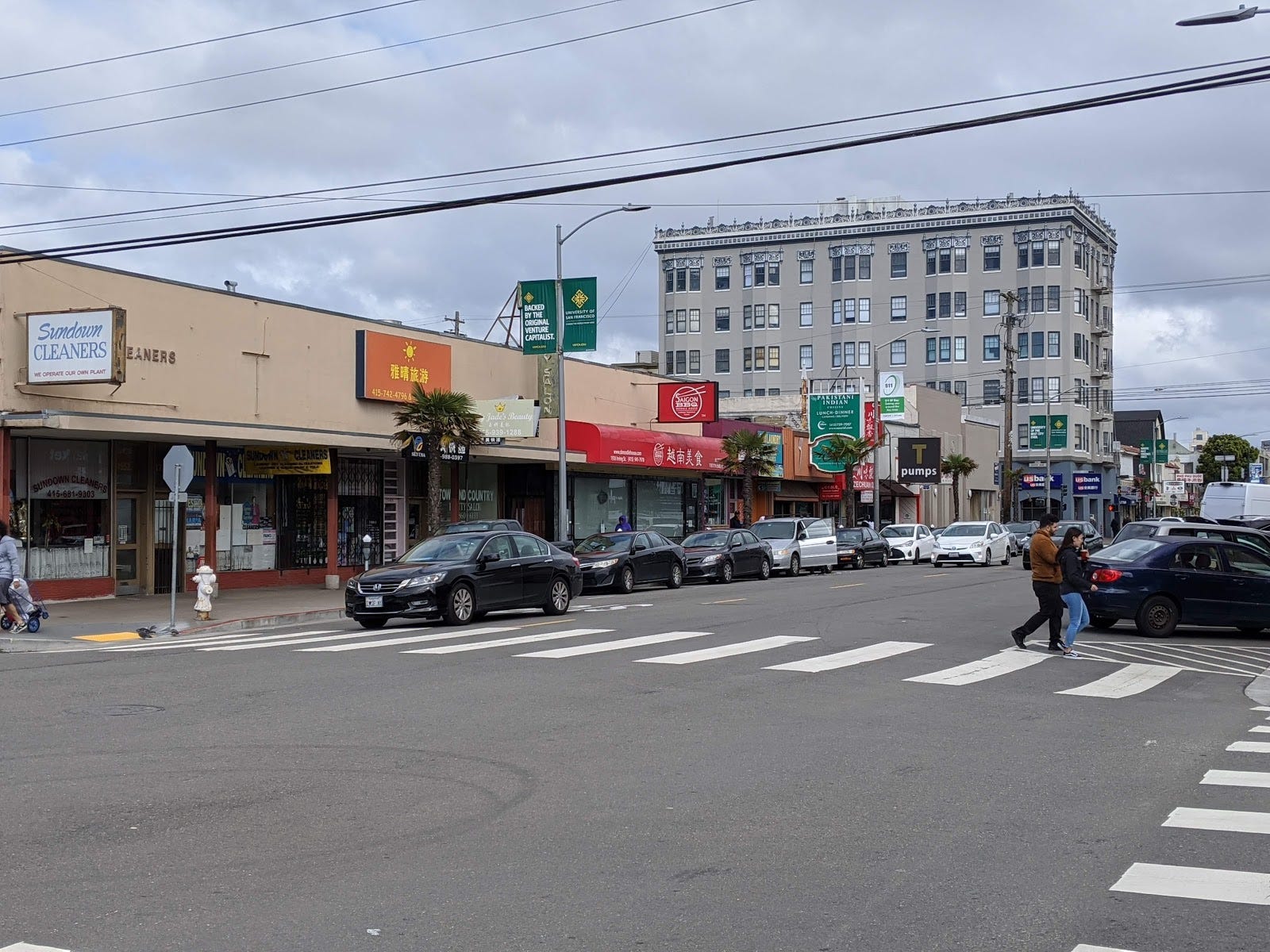 A view of the same street as above, but taken from the other side. A large apartment building is behind a row of shops.