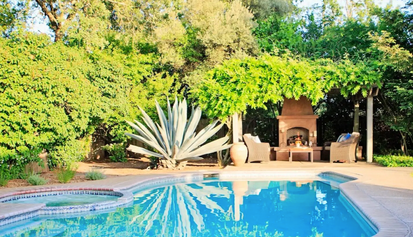 A pool with a hot tub, with trees, a large agave plant, and a brick fireplace with a sitting area in the background.