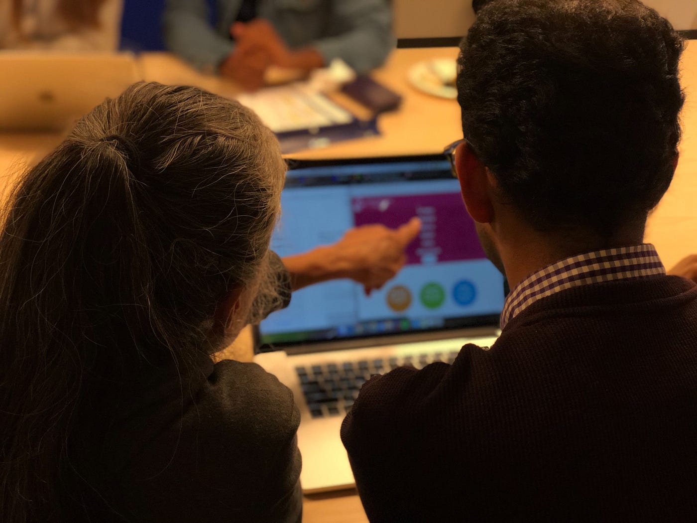Two people working at a a laptop, seen from behind. The person on the left is pointing at the screen.
