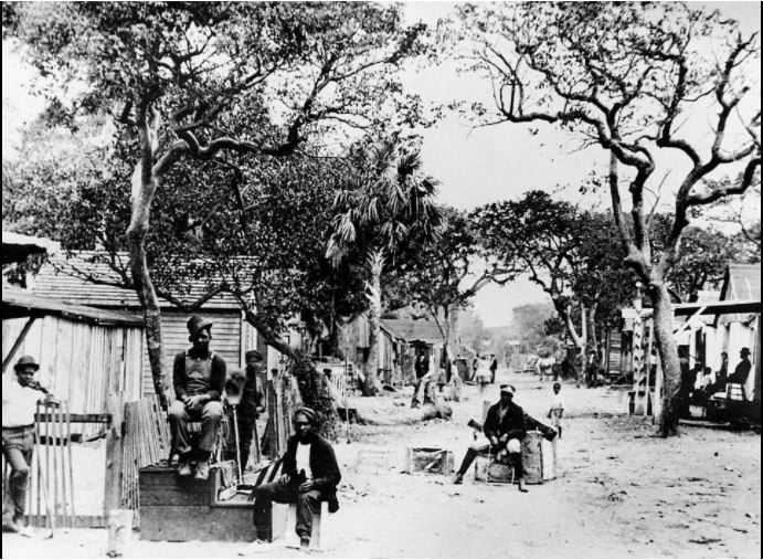 Black Americans pose for a photo on a dirt road in Florida's "The Styx" around 1900. The dirt road is line with shacks that appear to be the homes of the people in the photo.