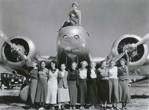 Earhart sits atop her famous twiin engined Lockheed Electra looking at the camera. Ten women, presumably studetns, face the camera, arm in arm.