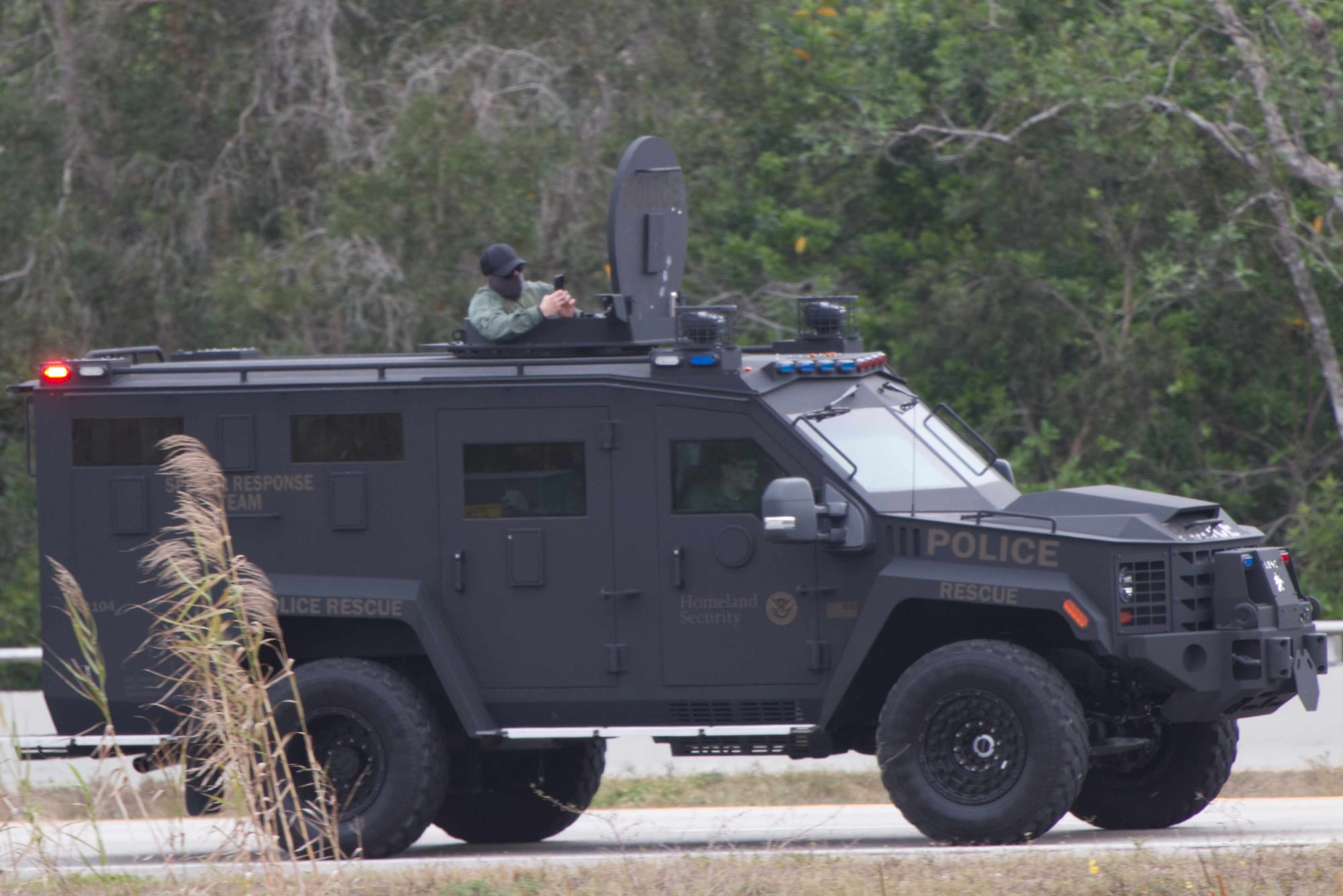 A homeland security armored personnel carrier driving with multiple officers using phones to take pictures of protestors. One officer is in the turret of the vehicle's roof and has his face entirely concealed.
